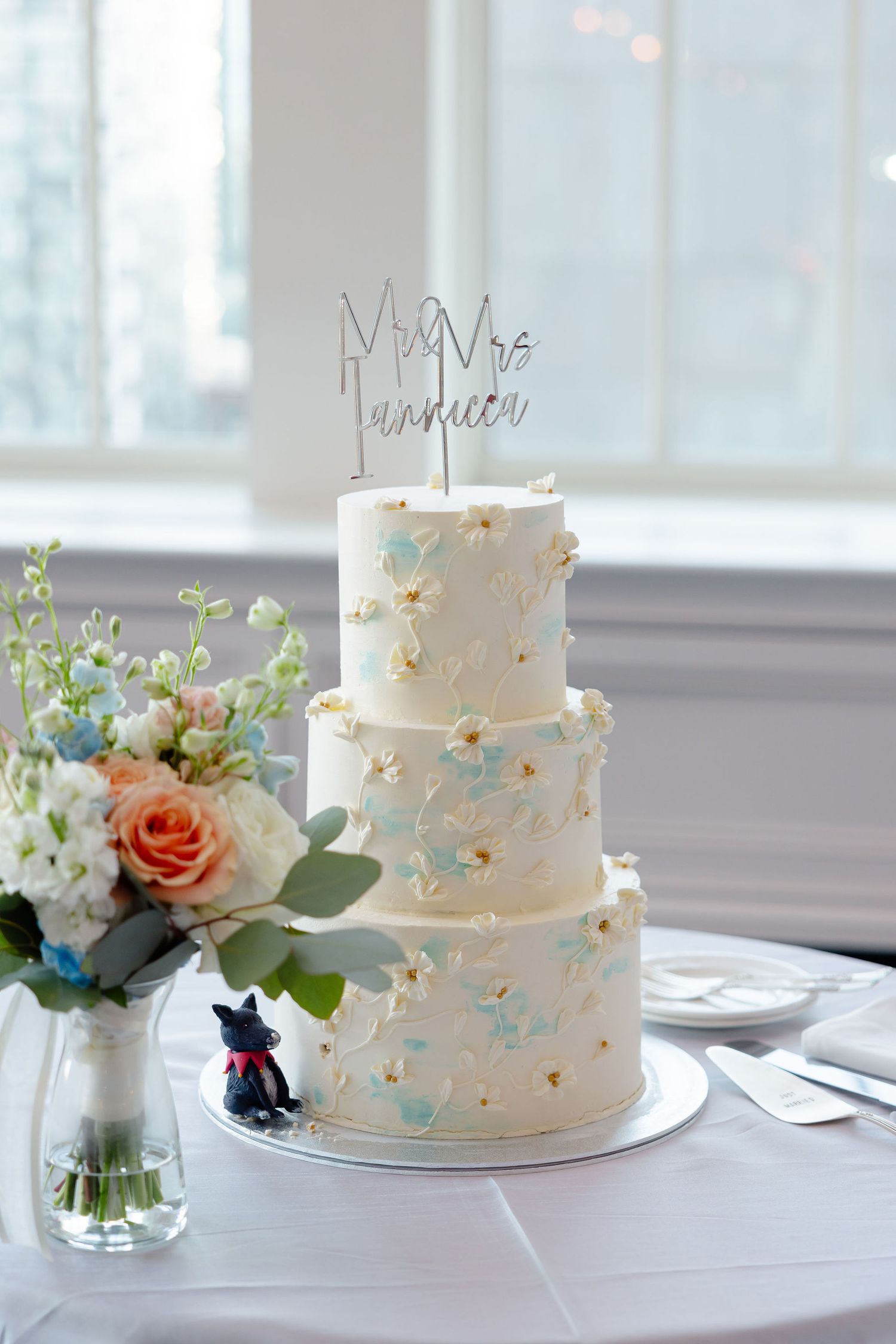 White wedding cake decorated with light blue accents and fresh flowers displayed by a window.