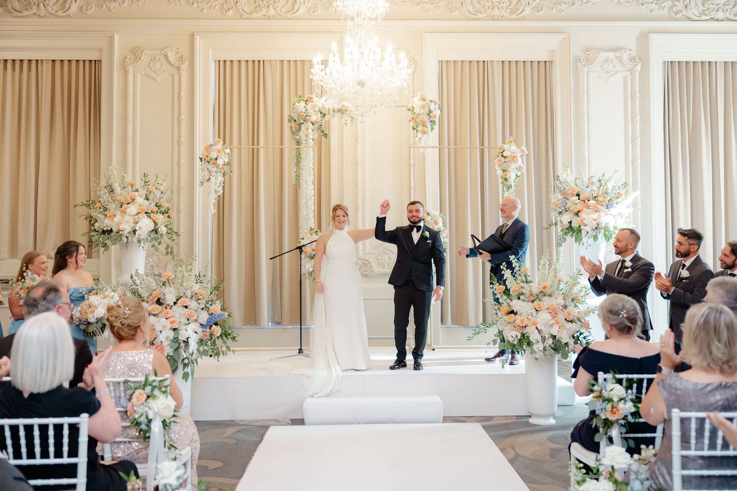 Wedding ceremony setup in a luxurious white ballroom with ornate moldings and crystal chandeliers.