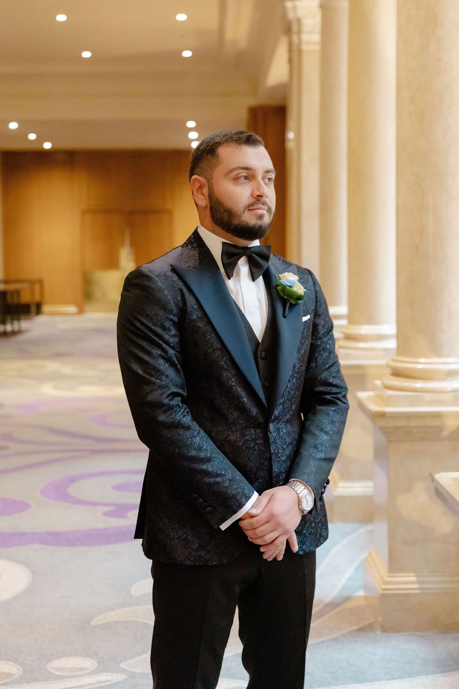 Groom in black tuxedo stands confidently in an ornate hallway with decorative floor patterns and columns.