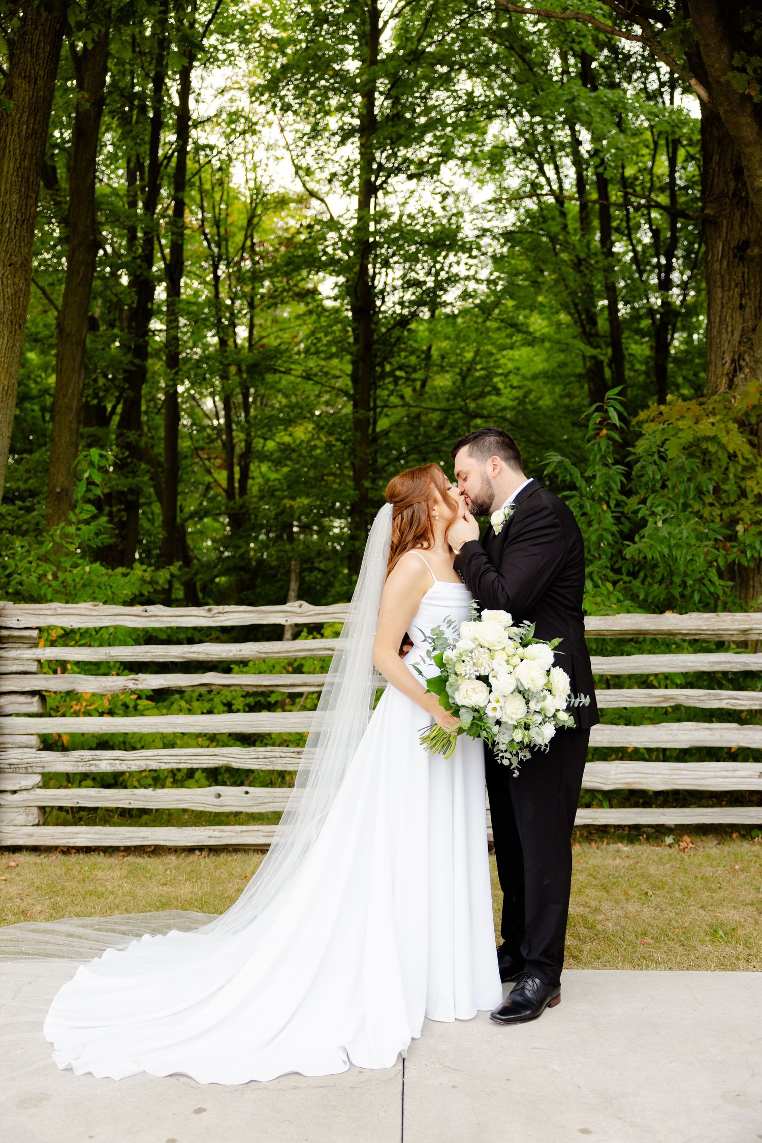 Full-length wedding portrait of couple embracing in front of wooden fence with forest background.