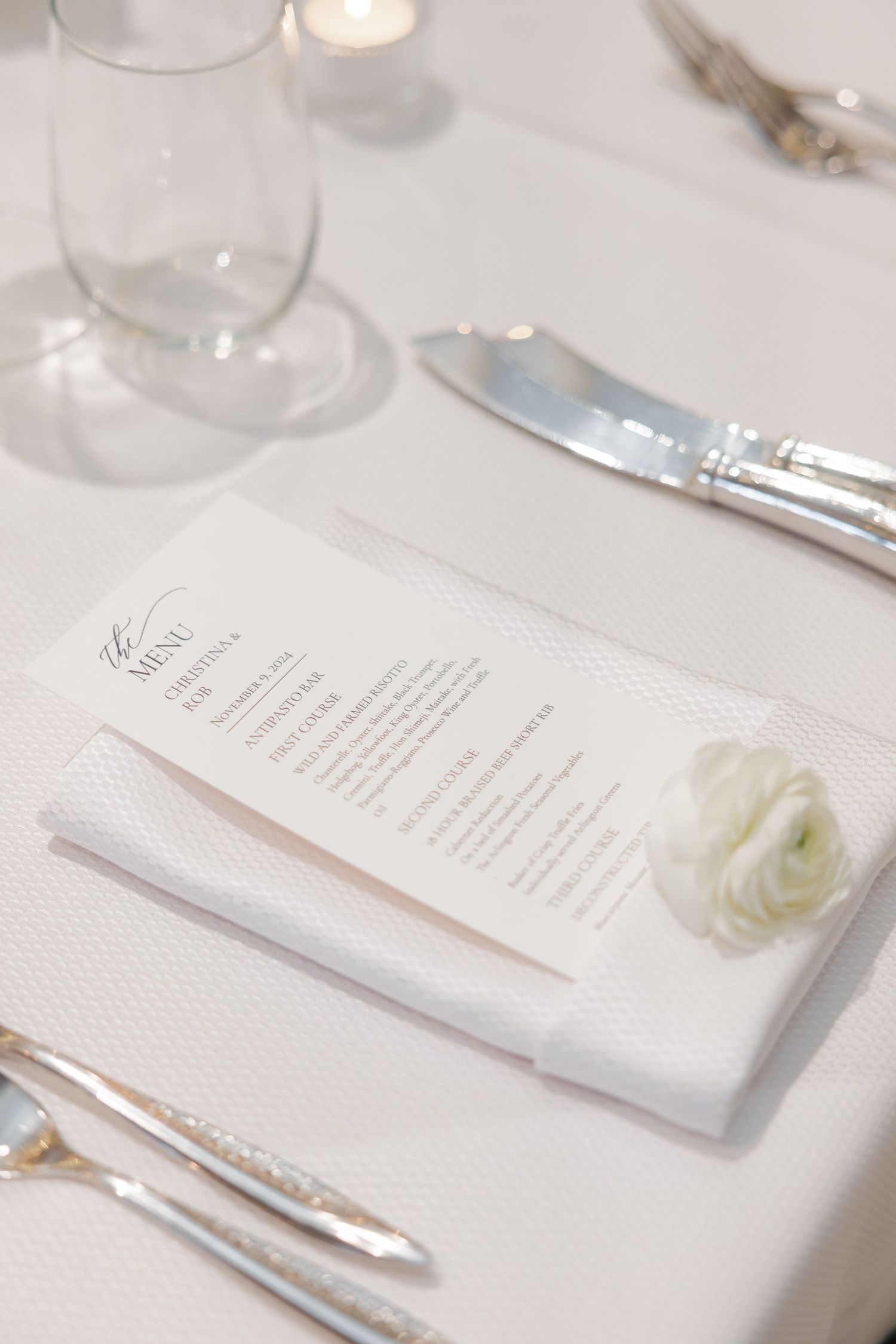 Wedding place setting with menu card, white napkin, and delicate white flower on a pristine tablecloth.