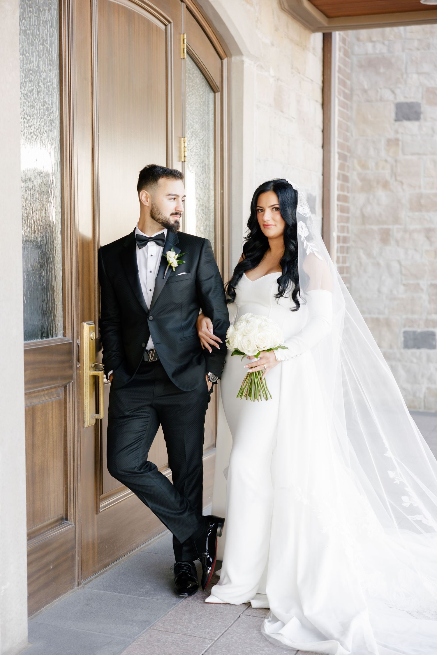 Elegant couple in formal wedding attire lean against door frame at stone venue.
