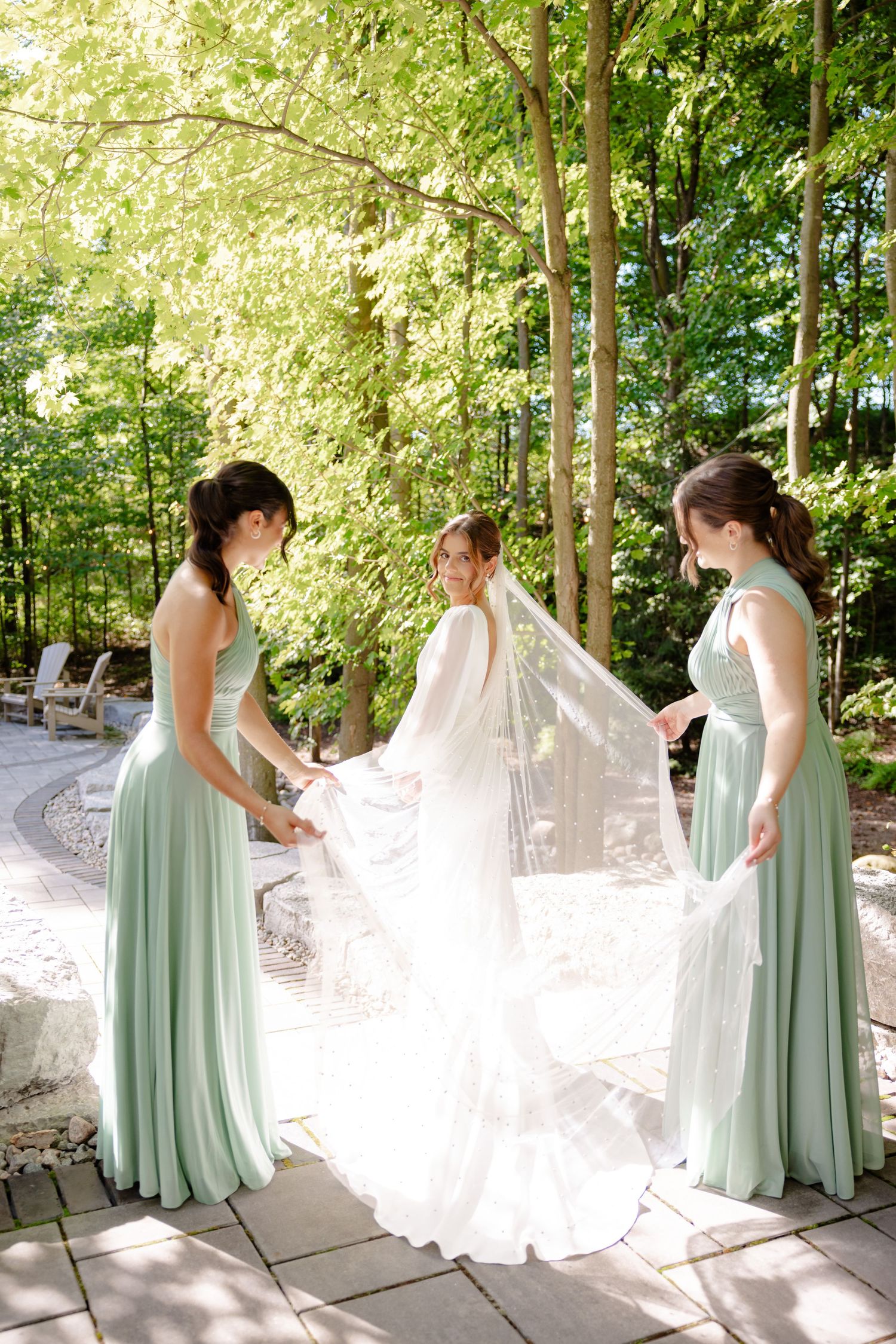Bridesmaids in sage green dresses help arrange bride's flowing veil in sunlit garden setting.