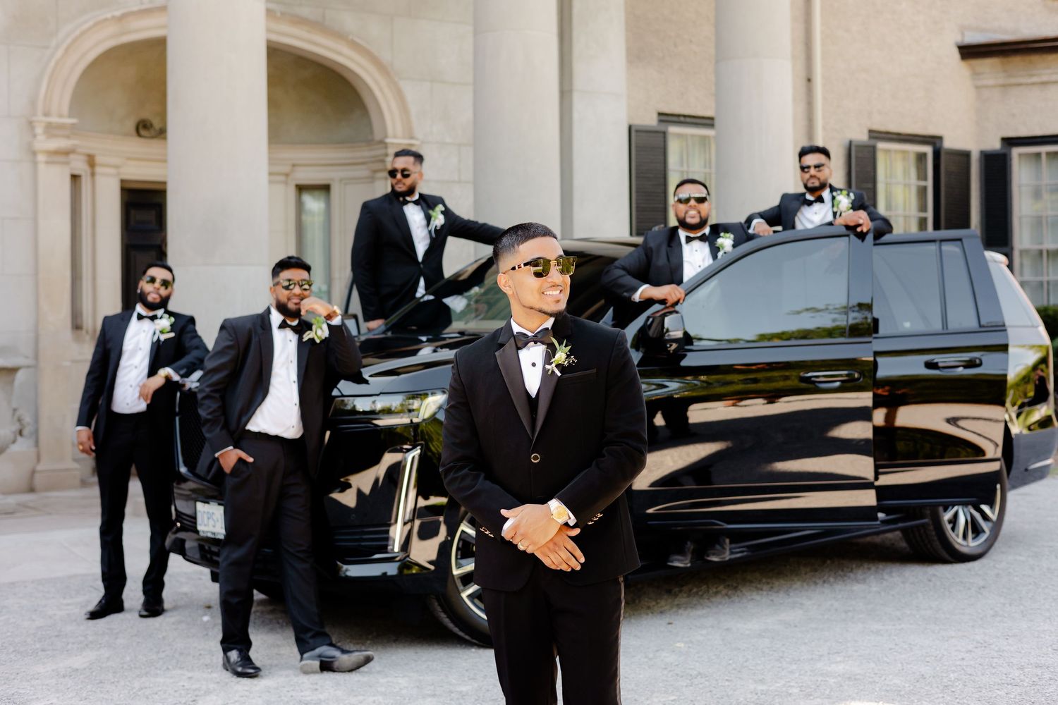 Wedding party members pose stylishly with a black luxury vehicle in front of a classic columned building.