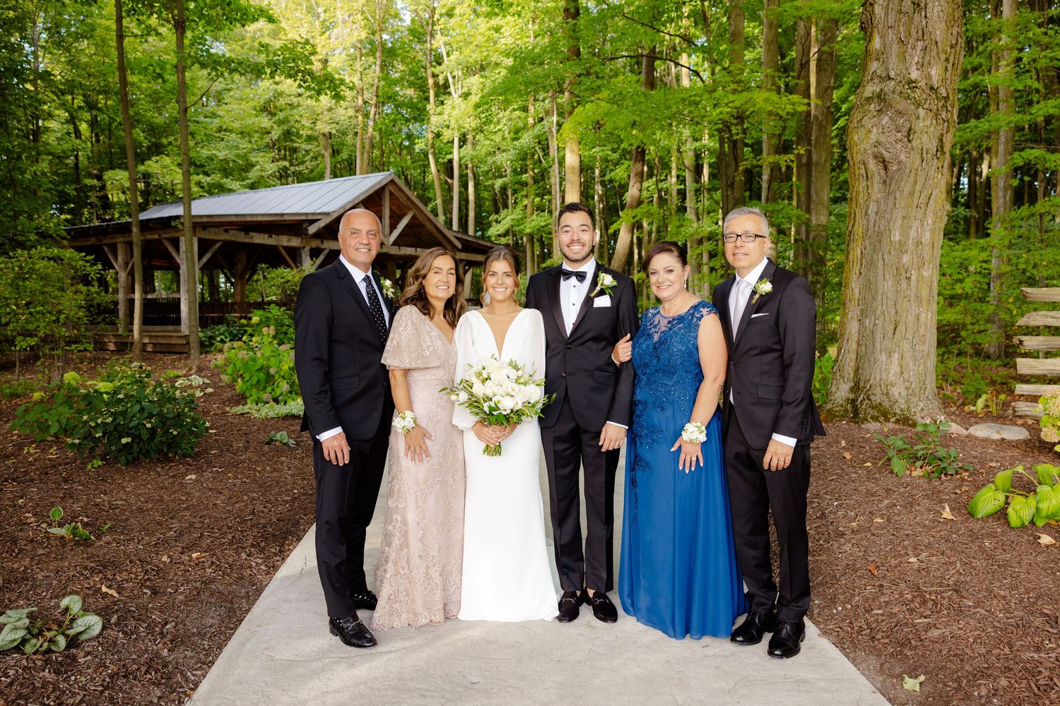 Wedding party poses on garden path in front of rustic pavilion surrounded by lush forest greenery.
