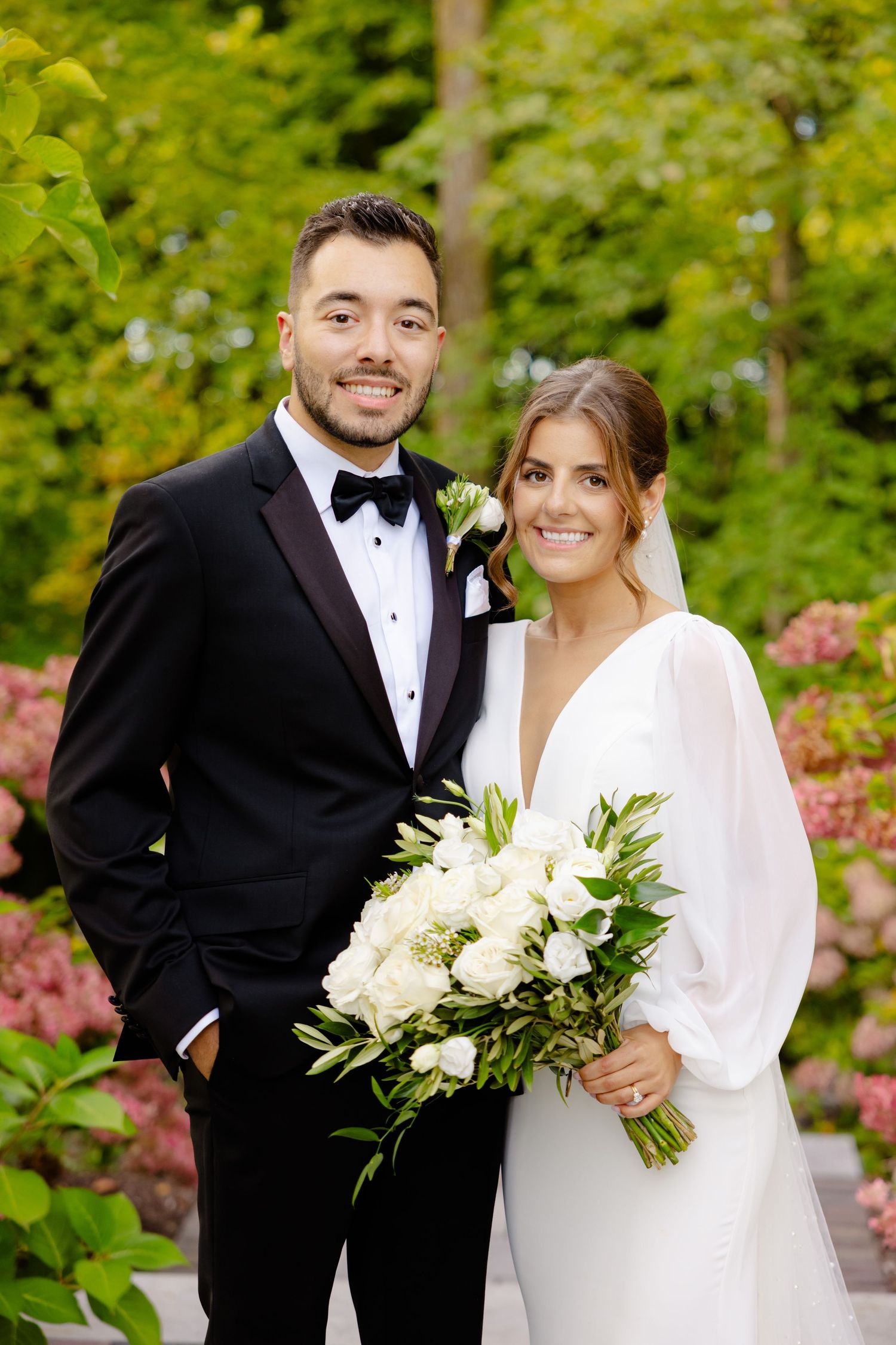 A newlywed couple poses in formal attire with a white floral bouquet against a garden backdrop.