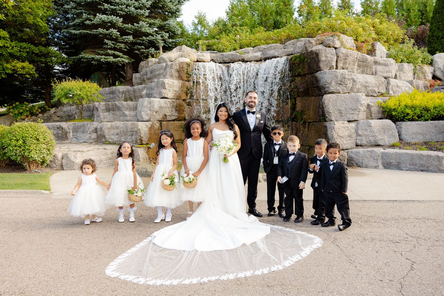 A wedding party poses in front of a stone waterfall backdrop with flower girls and ring bearers.