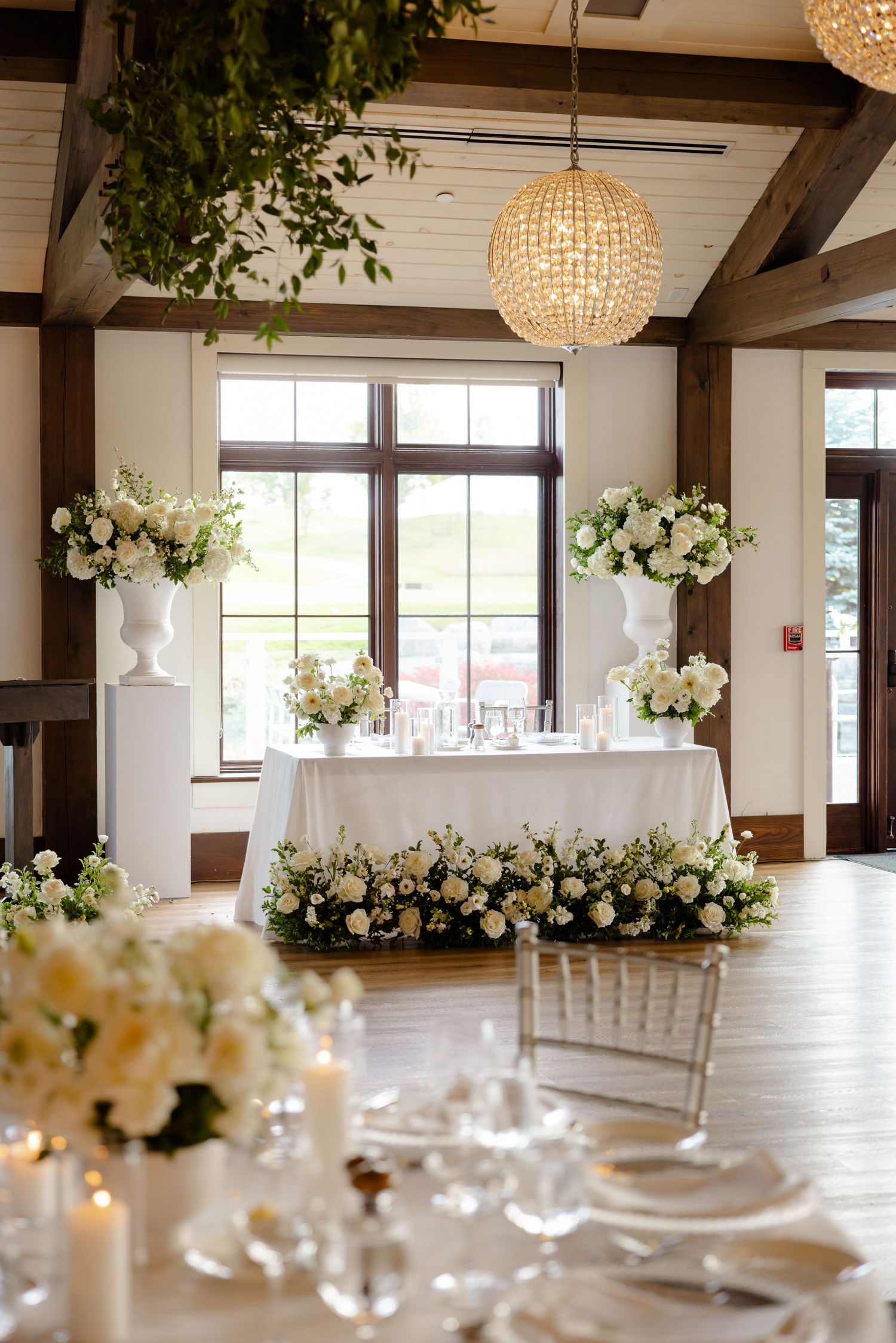 Wedding reception setup with white floral centerpieces, crystal lighting and wooden beam architecture.
