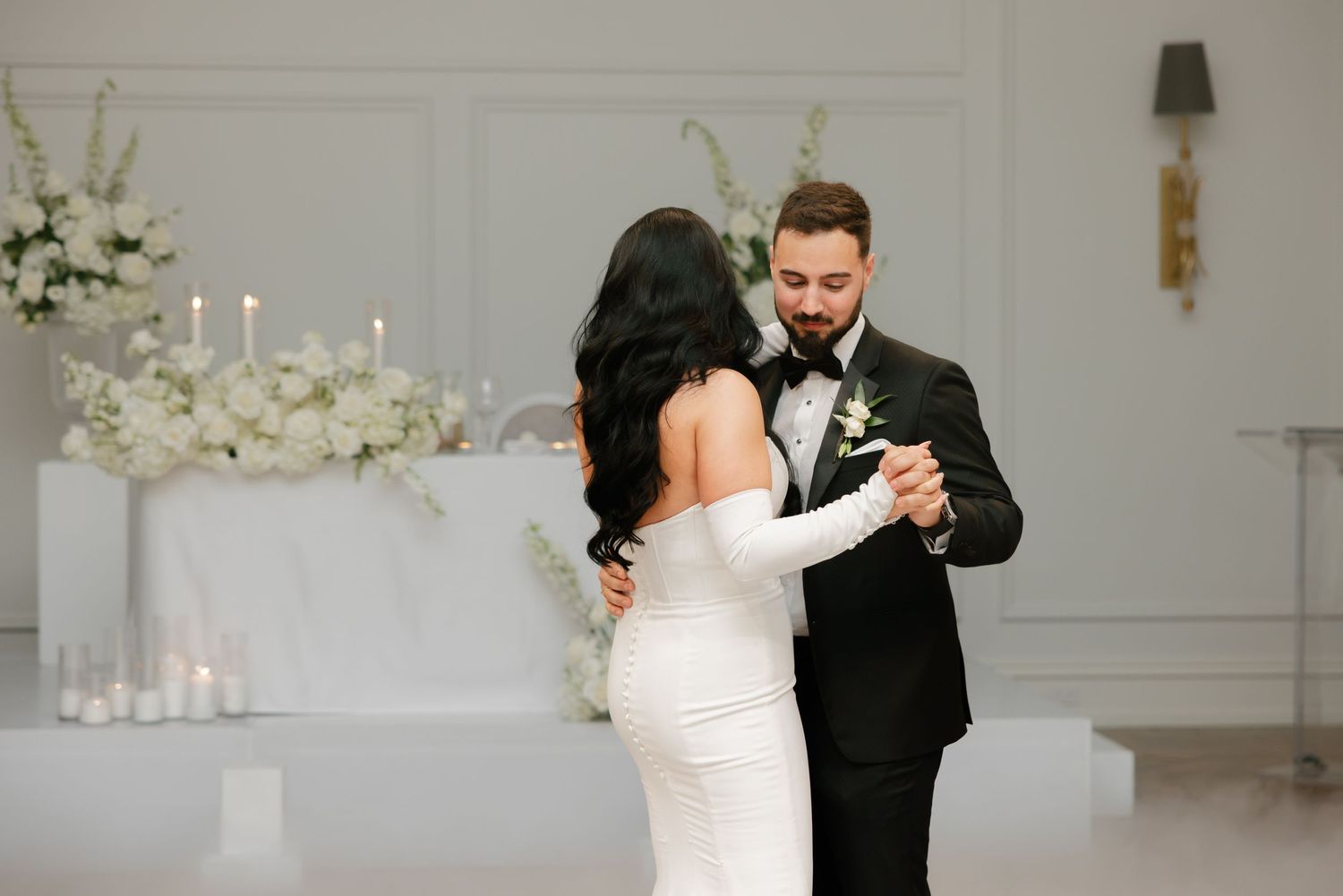 Couple dances in elegant ballroom with white floral arrangements and candles in background.