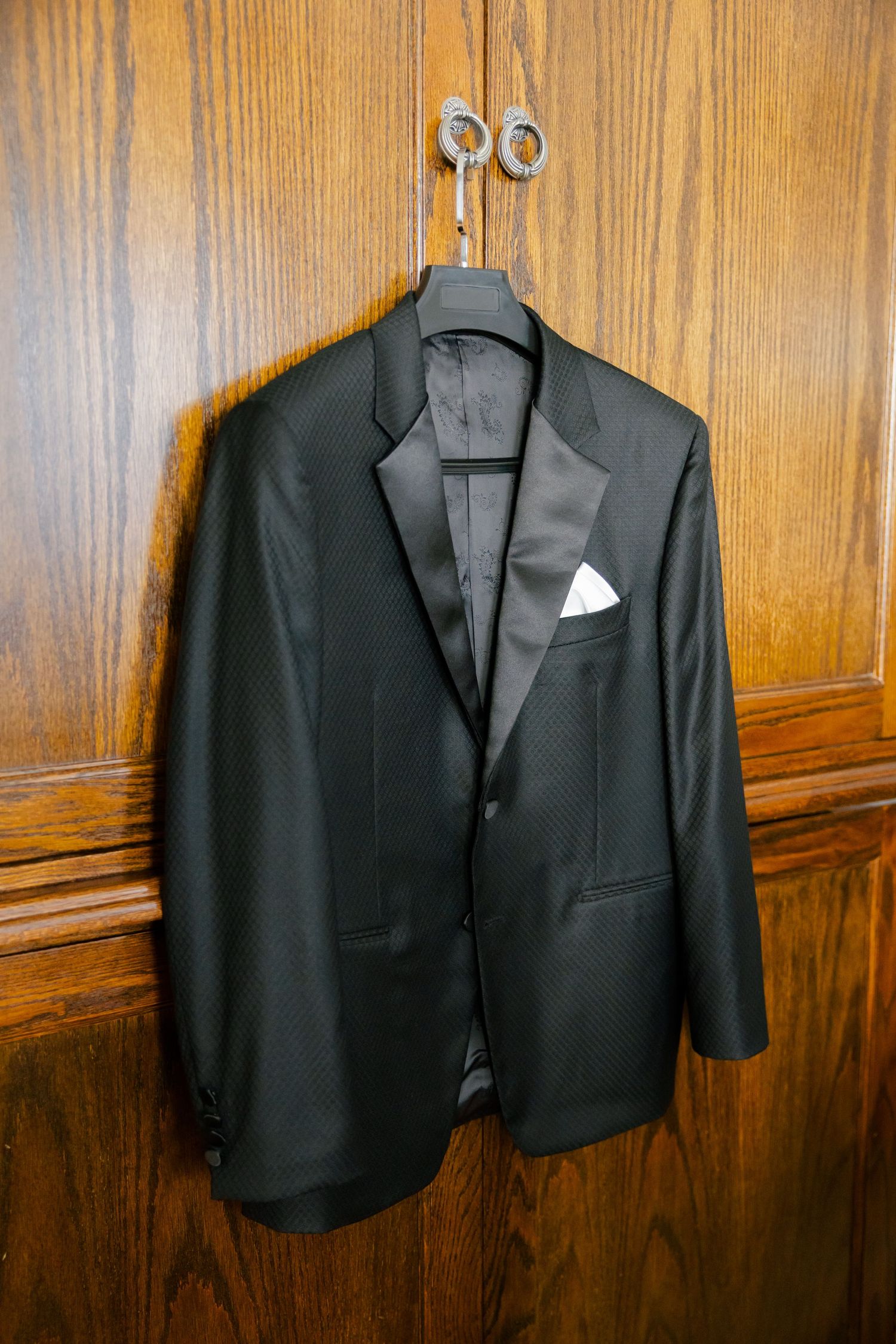 Classic black tuxedo jacket with white pocket square hanging on wooden wardrobe.