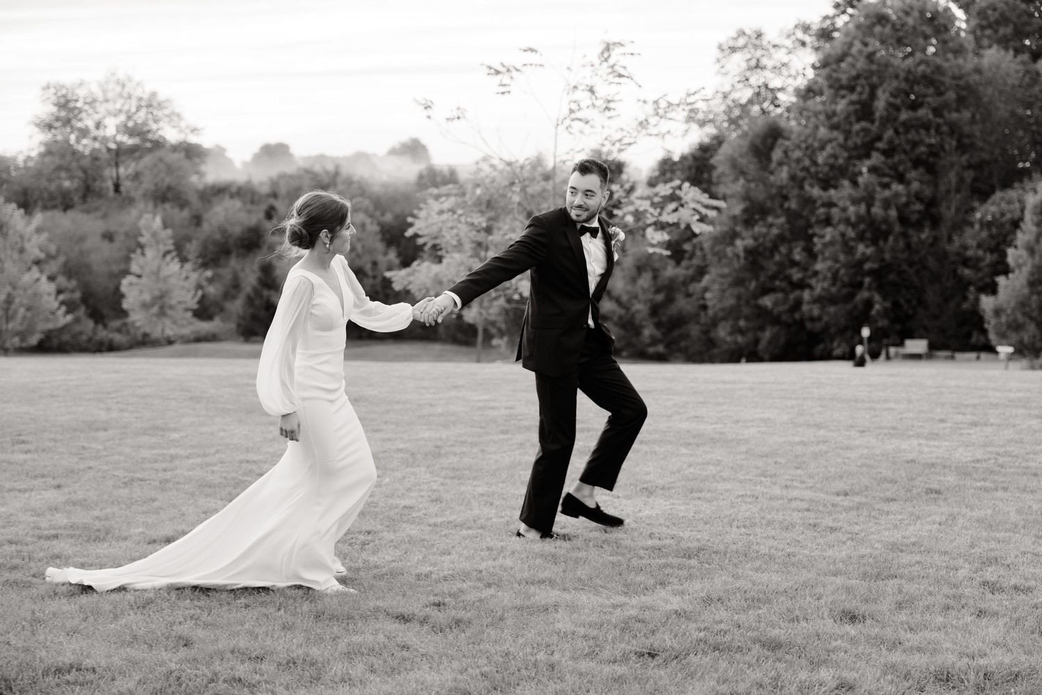 A playful black and white photo of a couple dancing in a field during their wedding.