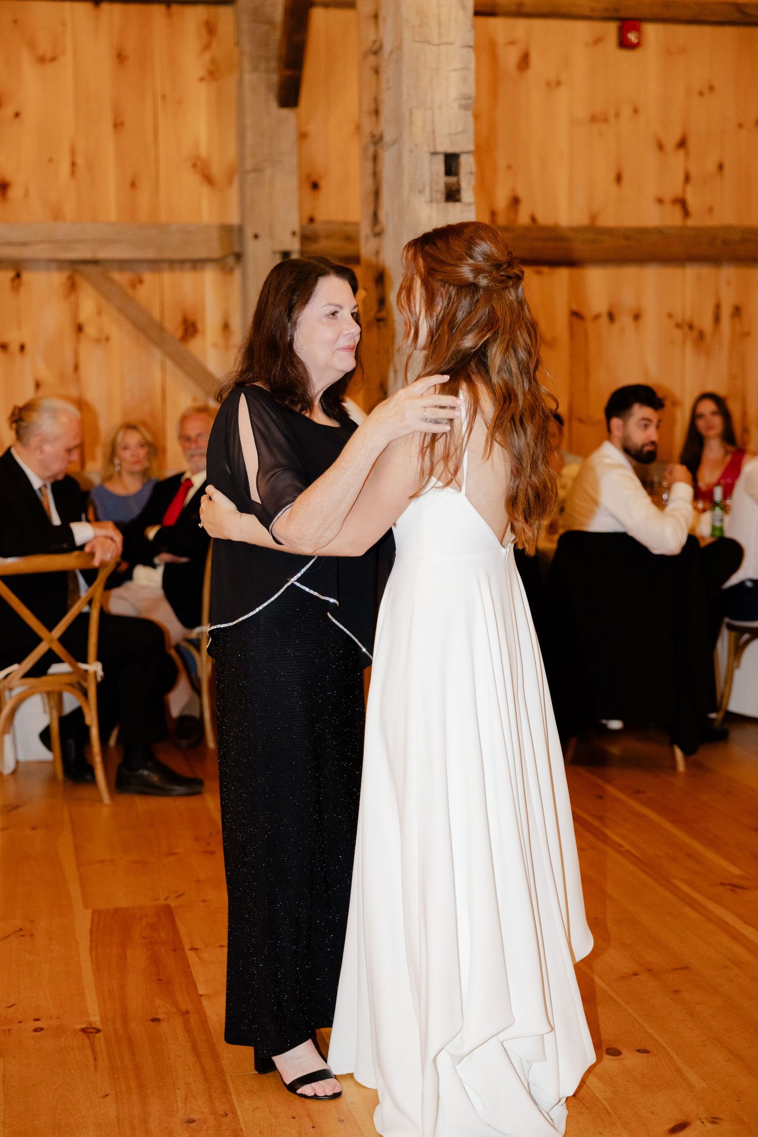 Two people share an emotional dance moment at a barn wedding reception with guests in background.