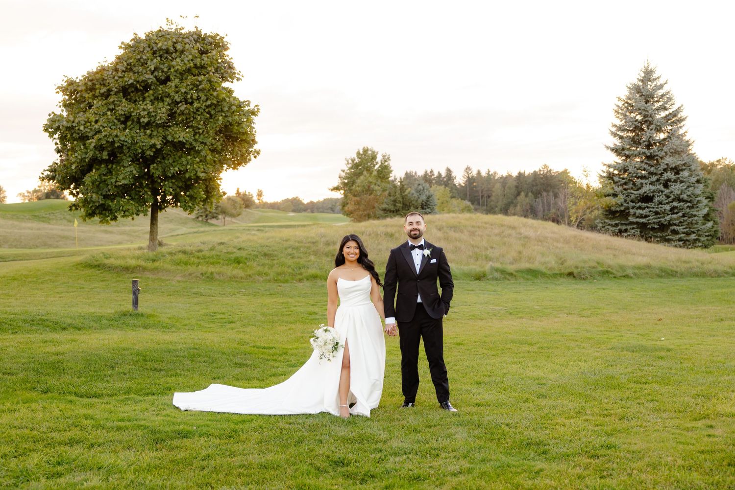 Wedding couple posing on rolling hillside with large tree and evergreens in background.