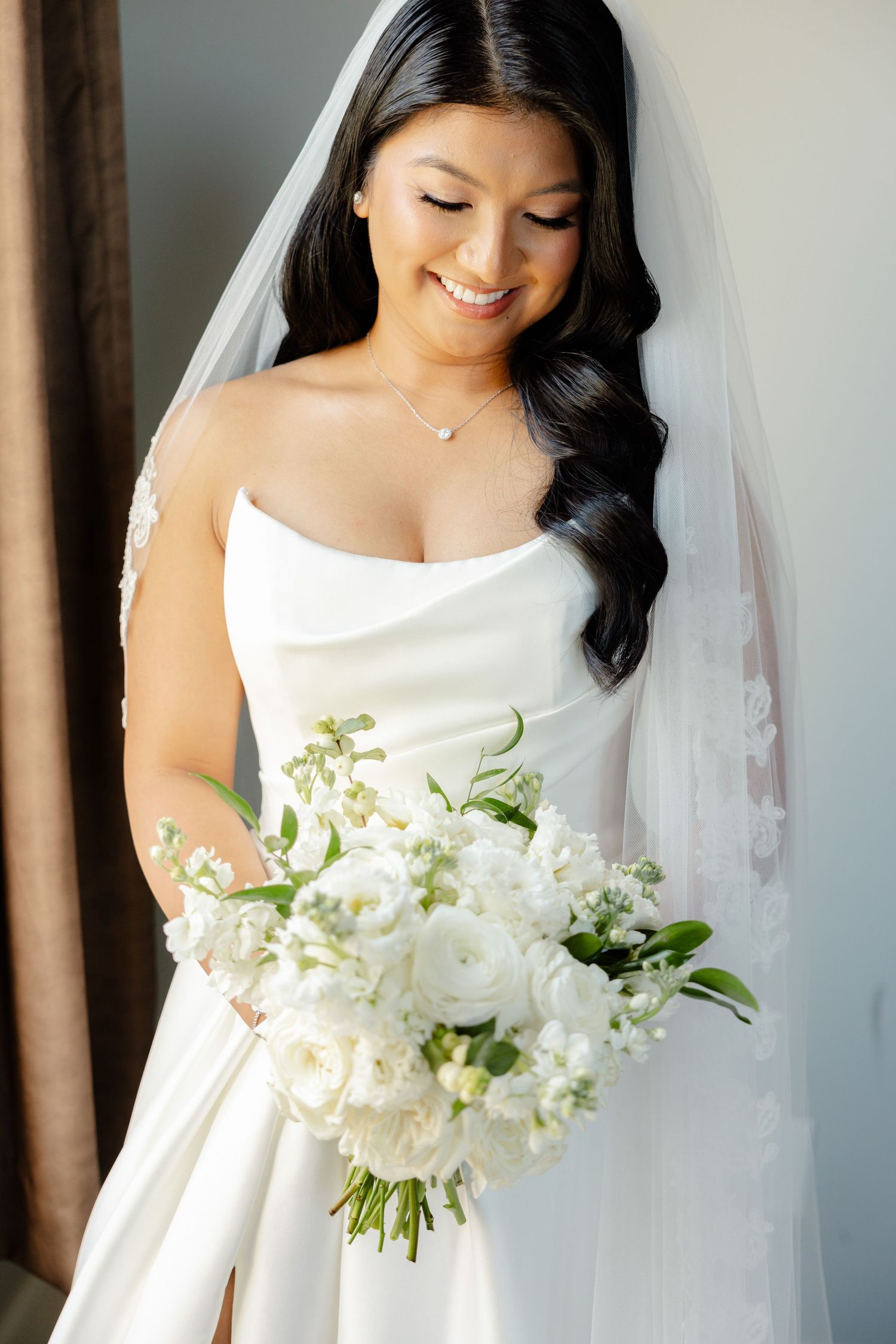 Bride in elegant white dress holding pure white bouquet with flowing veil and dark wavy hair styled to one side.