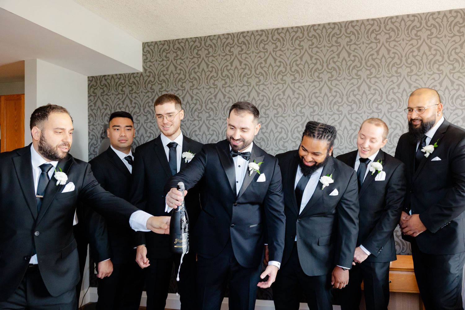A wedding party of groomsmen in black tuxedos and suits gathered in a room with decorative wallpaper.
