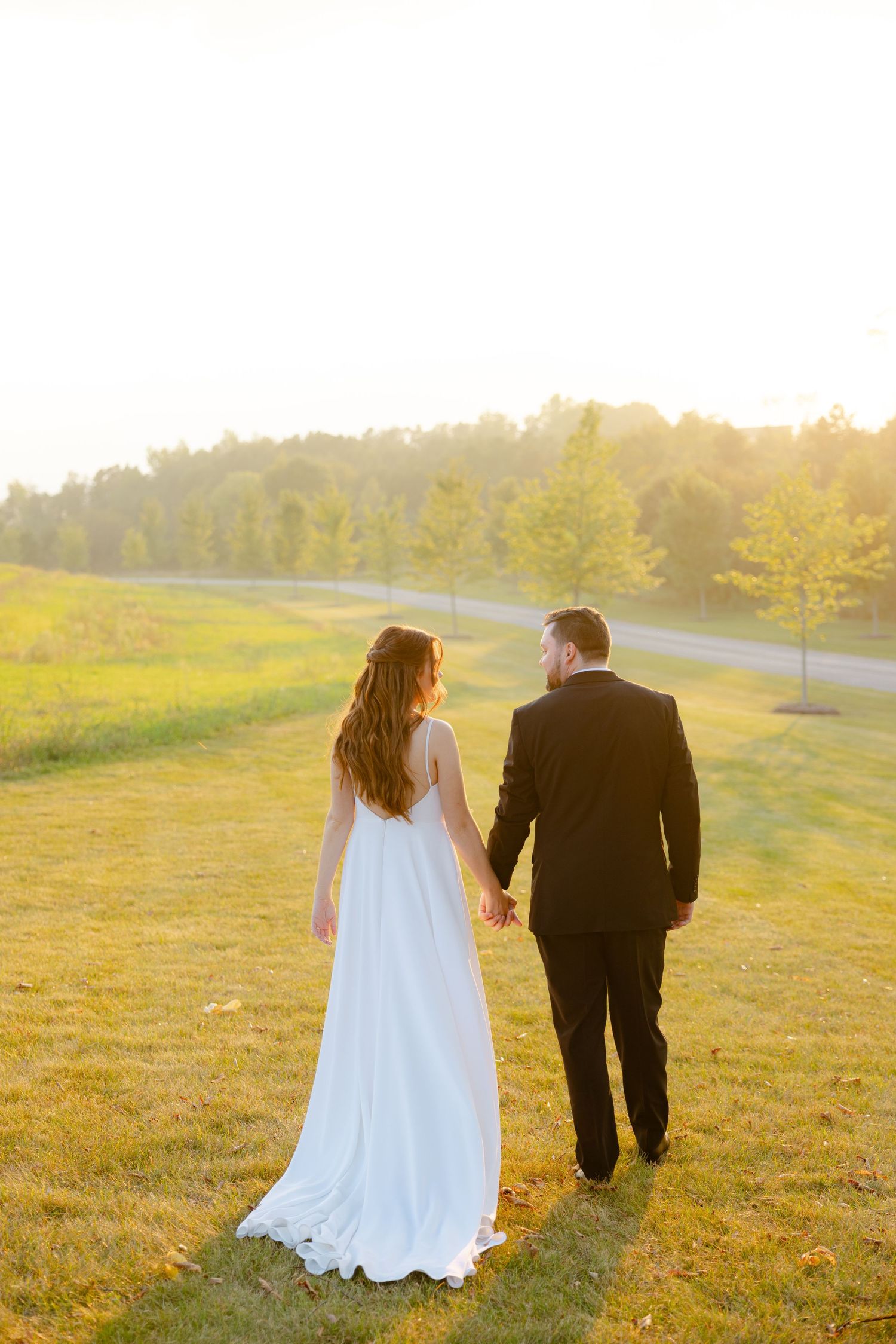 Newlyweds stroll together on a grassy hillside during golden hour with a winding road visible behind them.
