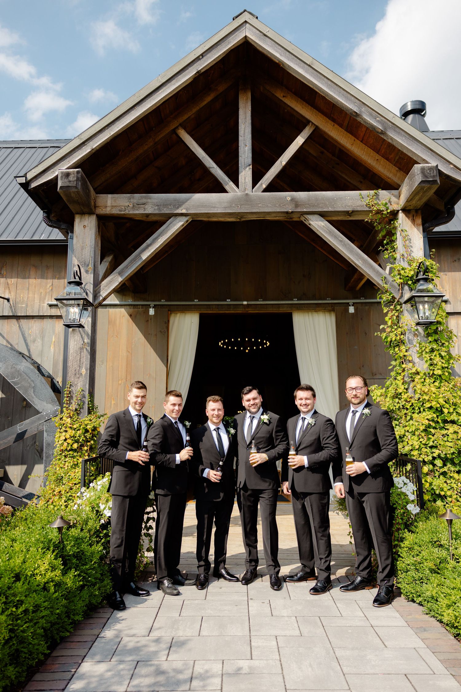 Wedding party poses in formal attire beneath a wooden beam entrance with climbing vines.