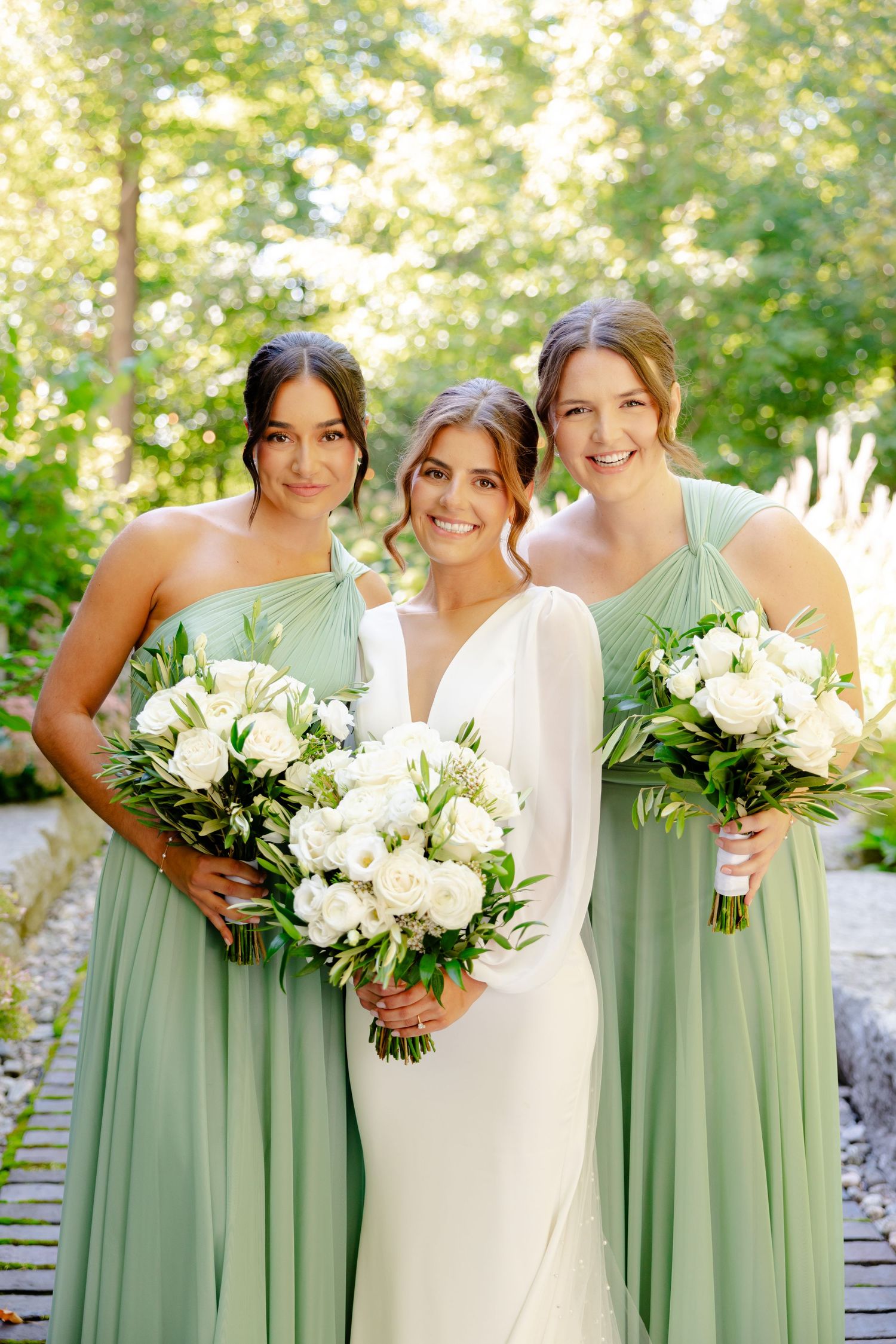Bridal party in sage green dresses and white wedding gown pose together with elegant white floral bouquets.