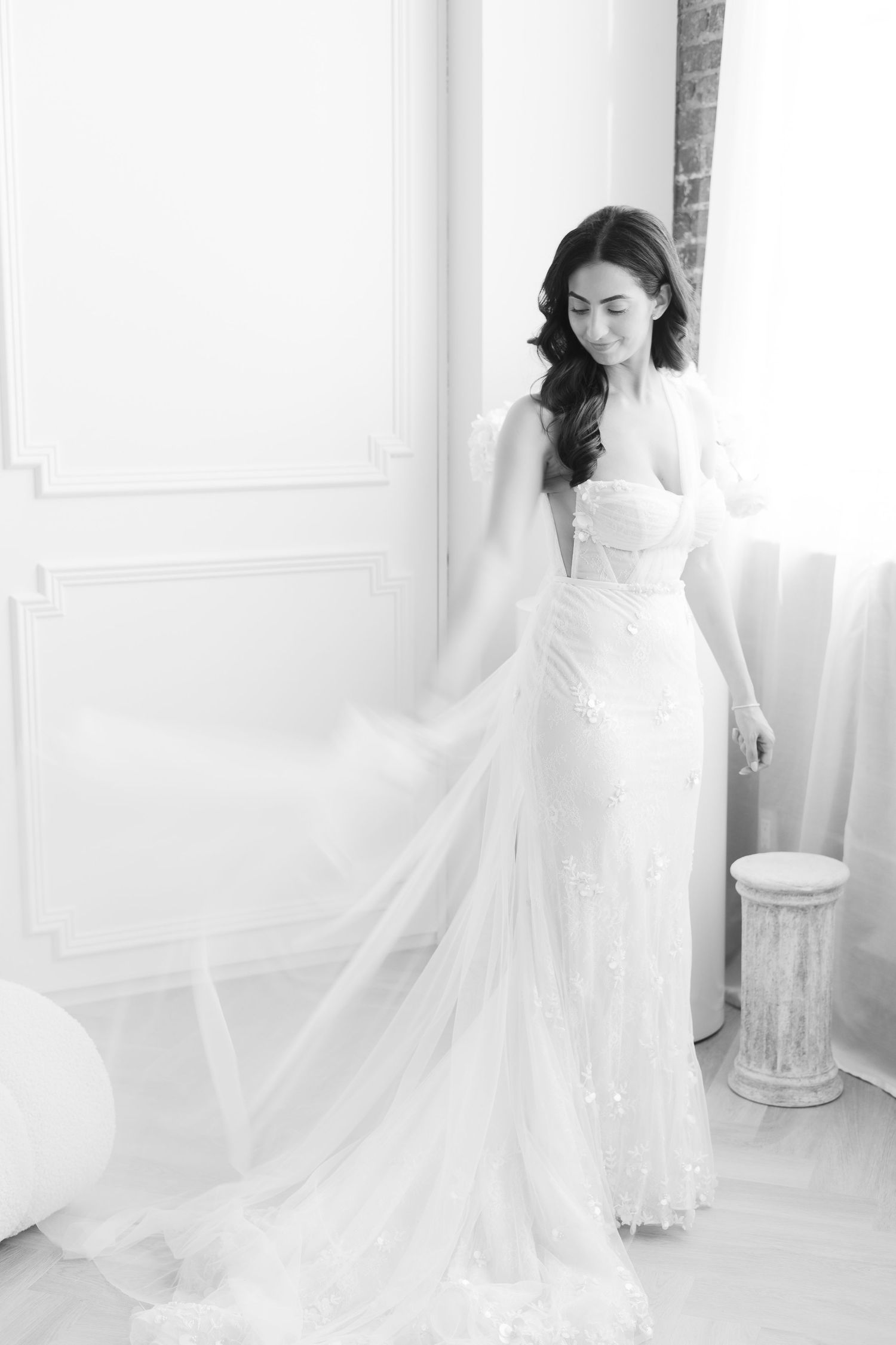 Black and white portrait of bride in flowing lace wedding dress by window.