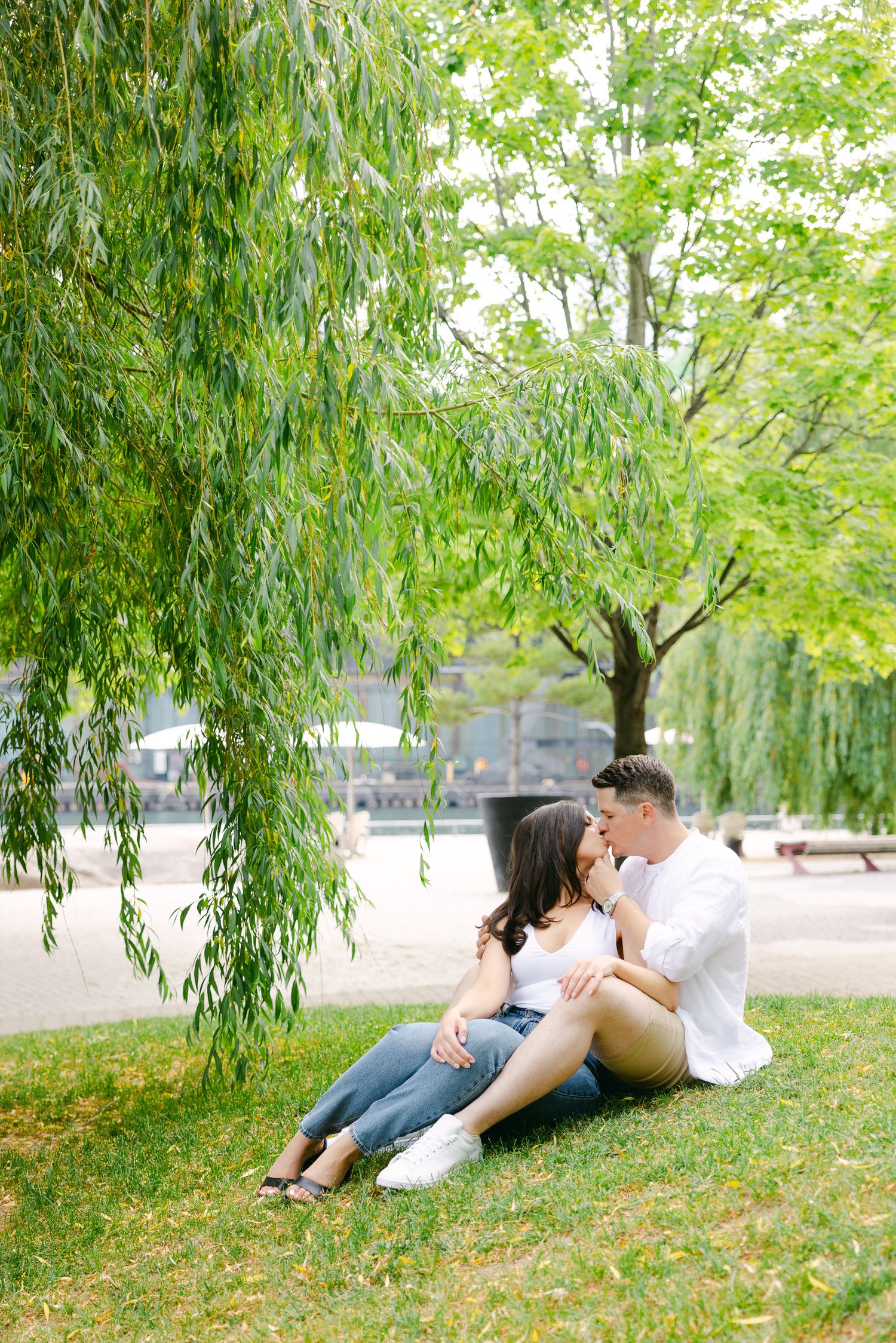Couple sharing tender moment while relaxing on grassy slope under willow tree.