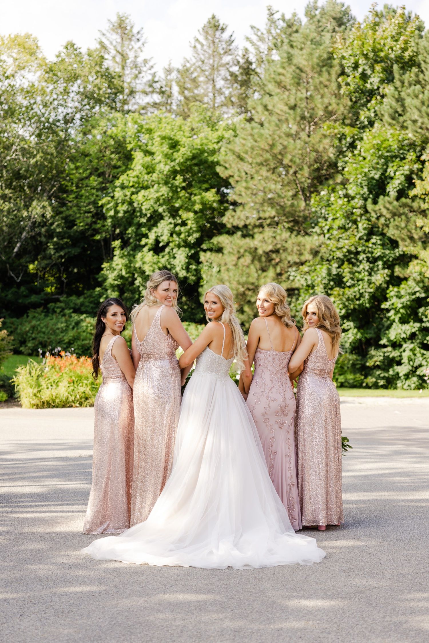 Bridal party in blush sequined dresses poses with bride in white gown against garden backdrop.