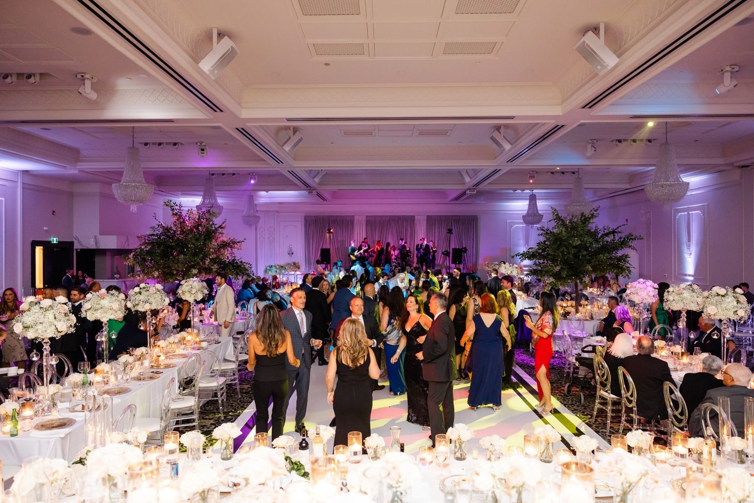 Wedding reception ballroom with purple lighting and guests mingling around decorated tables.