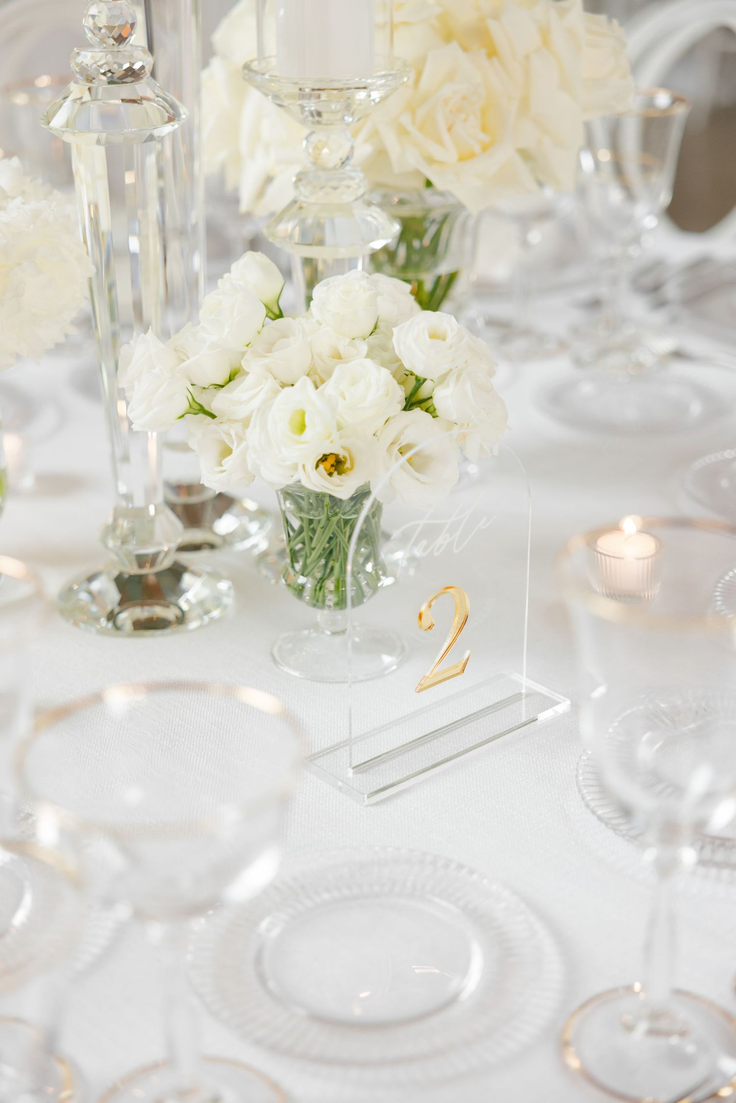 Elegant white wedding table setting with crystal vase of white roses and table number.