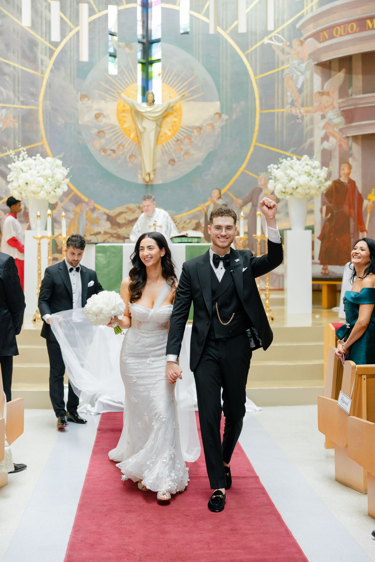 A wedding couple celebrates while walking down a church aisle decorated with white floral arrangements.