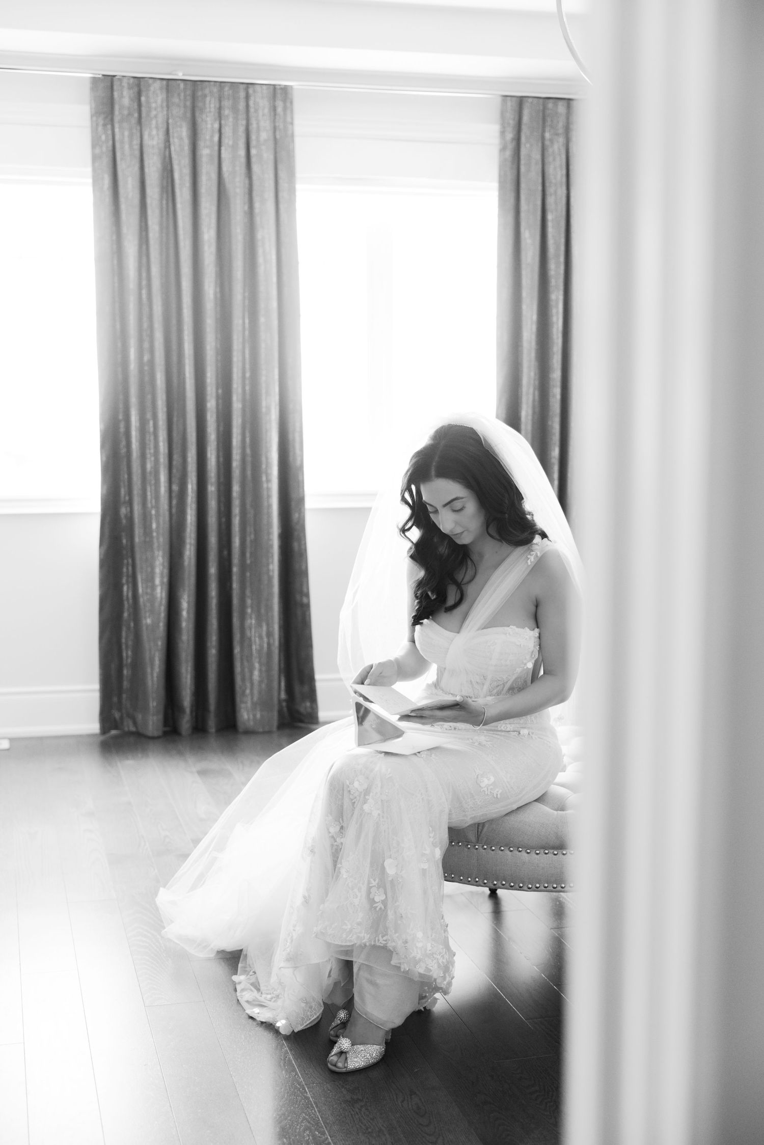 Bride in white gown sits reading a letter by window light in an intimate getting ready moment.