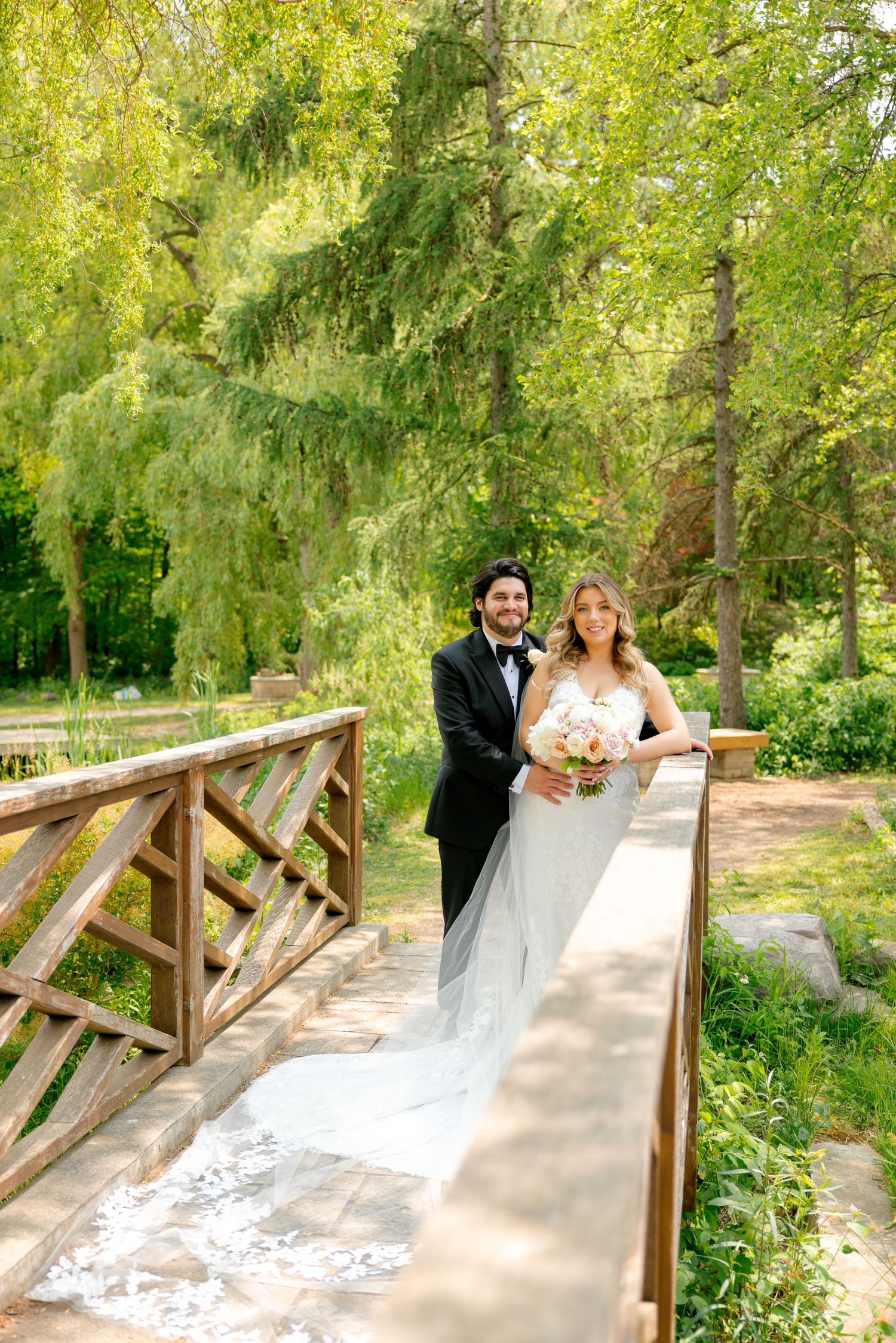 Joyful wedding portrait on wooden bridge with sunlit garden background.