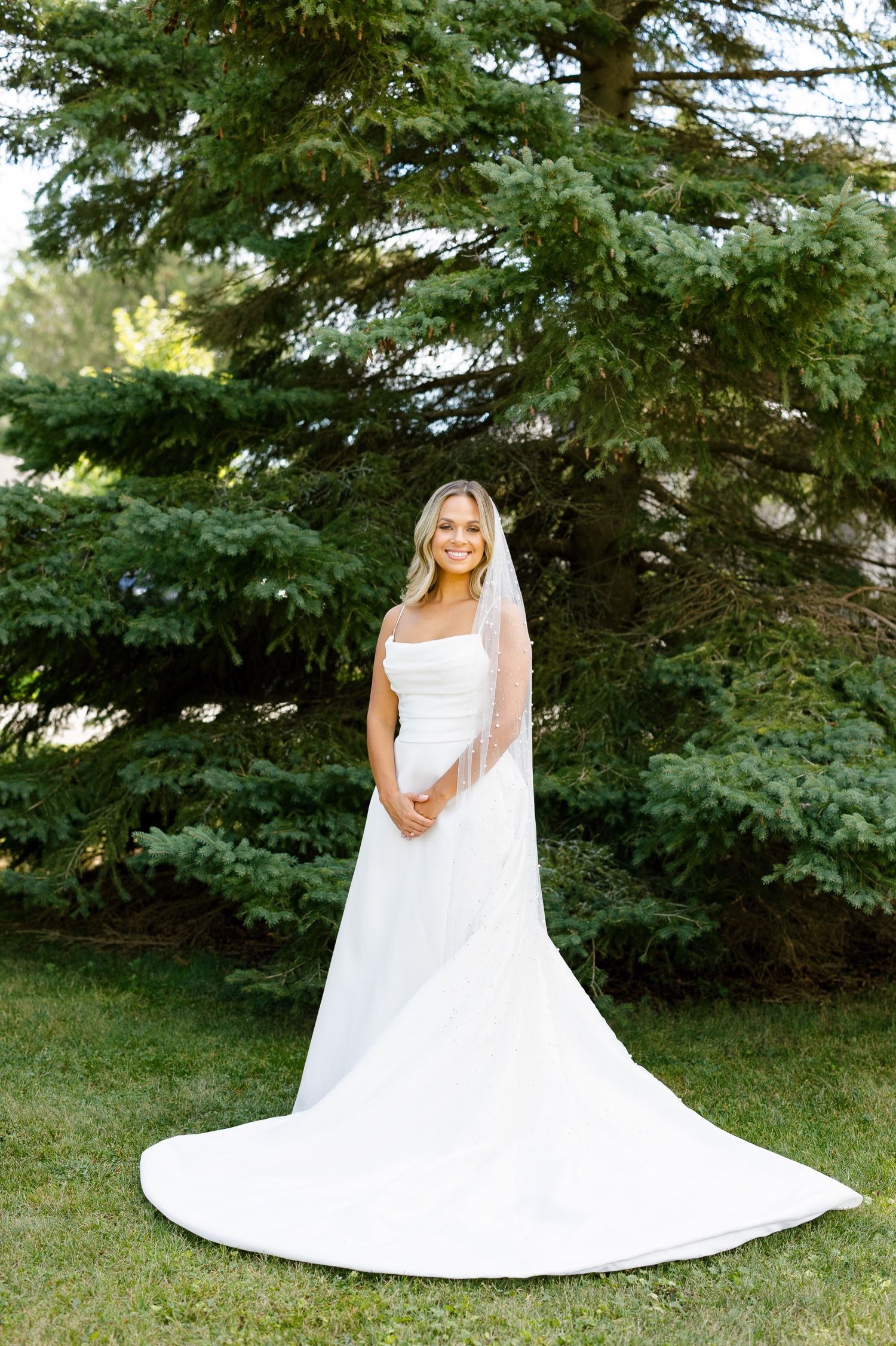 Bride in flowing white gown with train stands elegantly against evergreen tree backdrop.