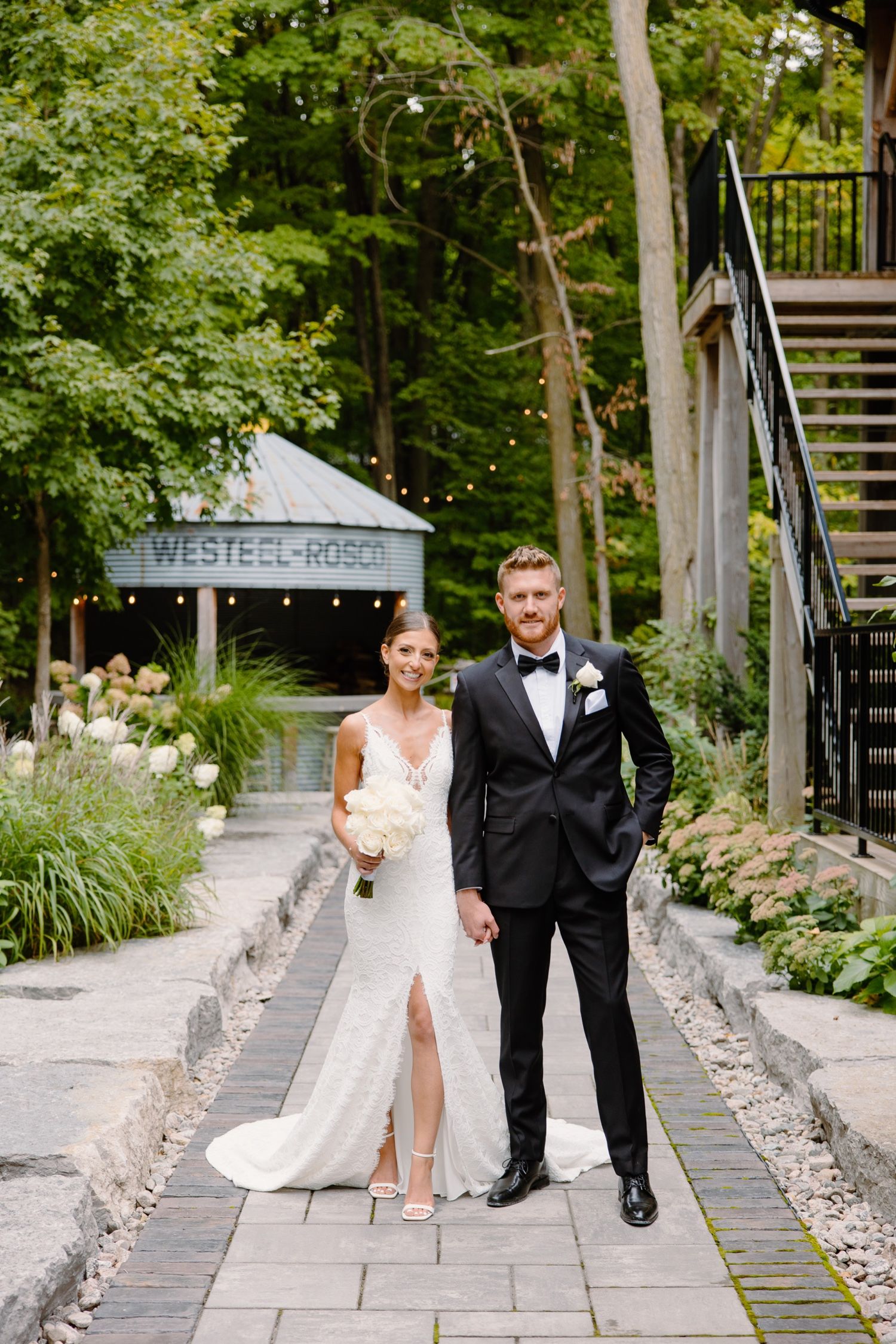 Elegant wedding couple poses on stone pathway with rustic venue and garden in background.