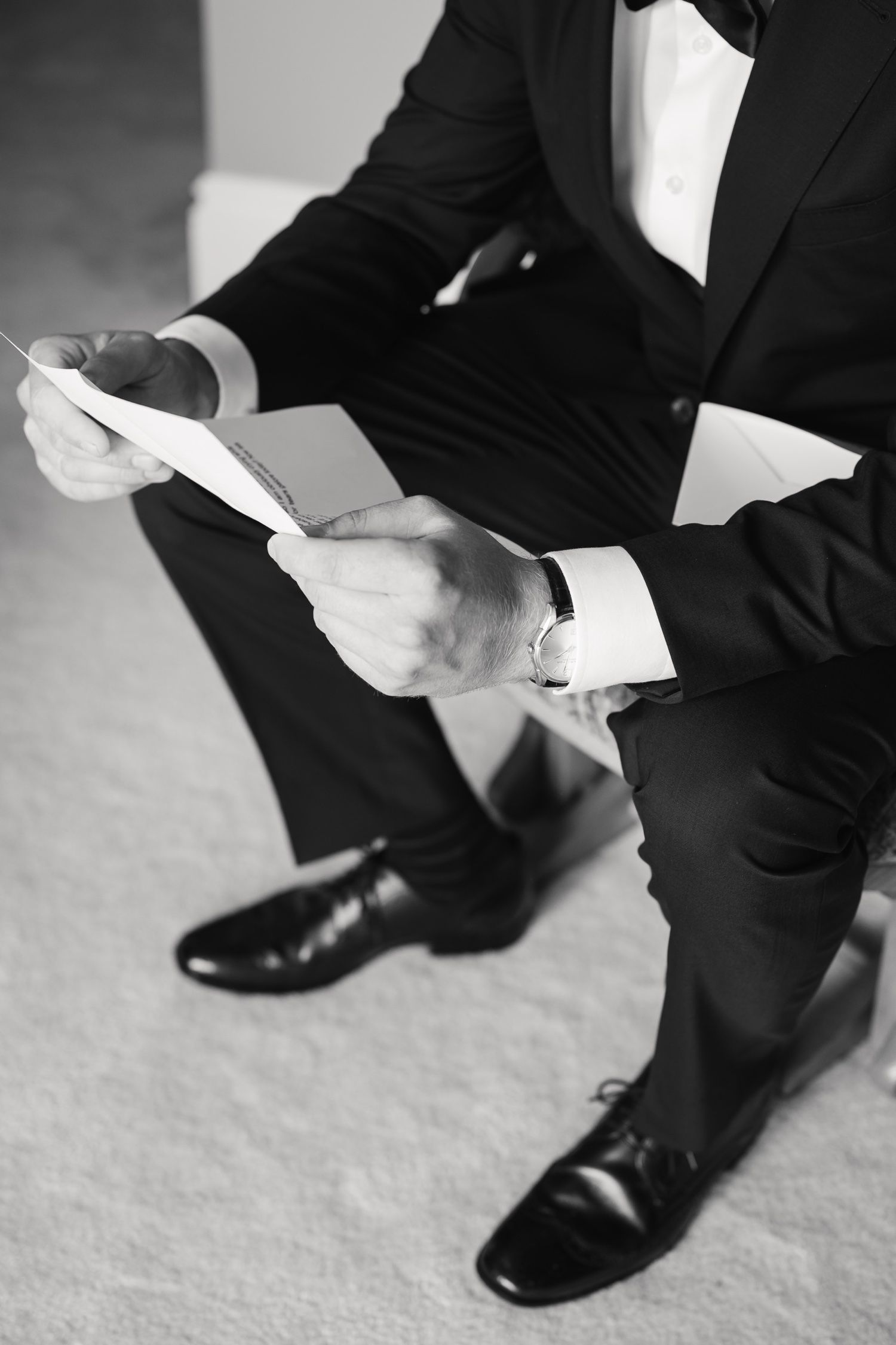Close-up of suited man in black dress shoes reading papers while sitting.