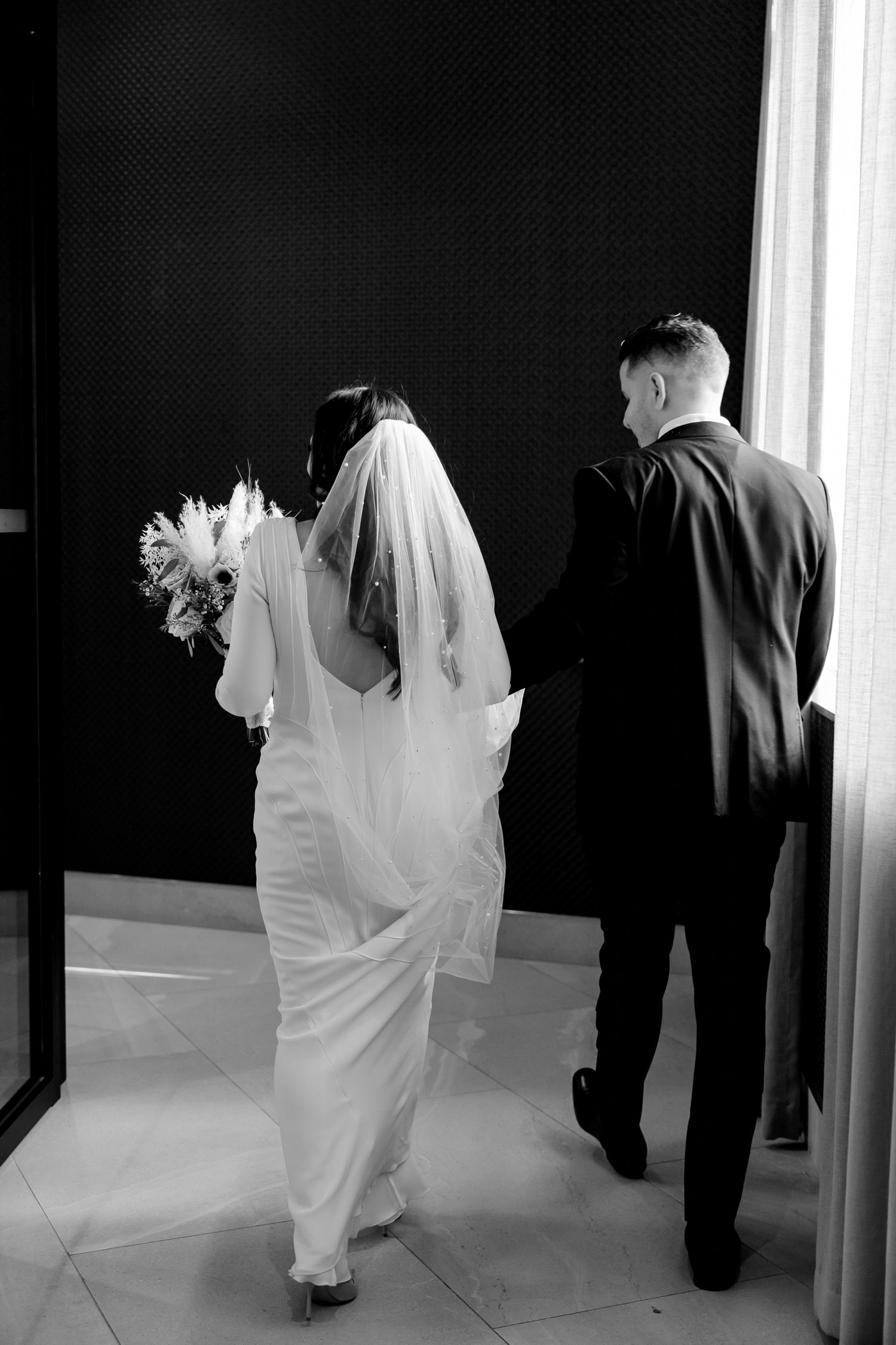 Black and white candid shot of newlyweds walking together, bride's elegant dress and veil trailing behind.