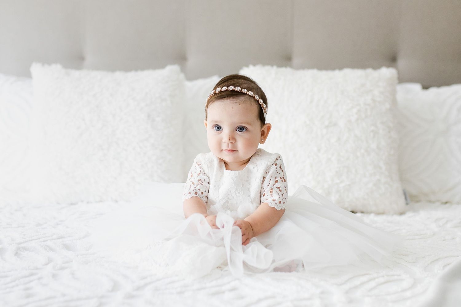 Baby wearing delicate white dress and headband sits among white pillows on bed.