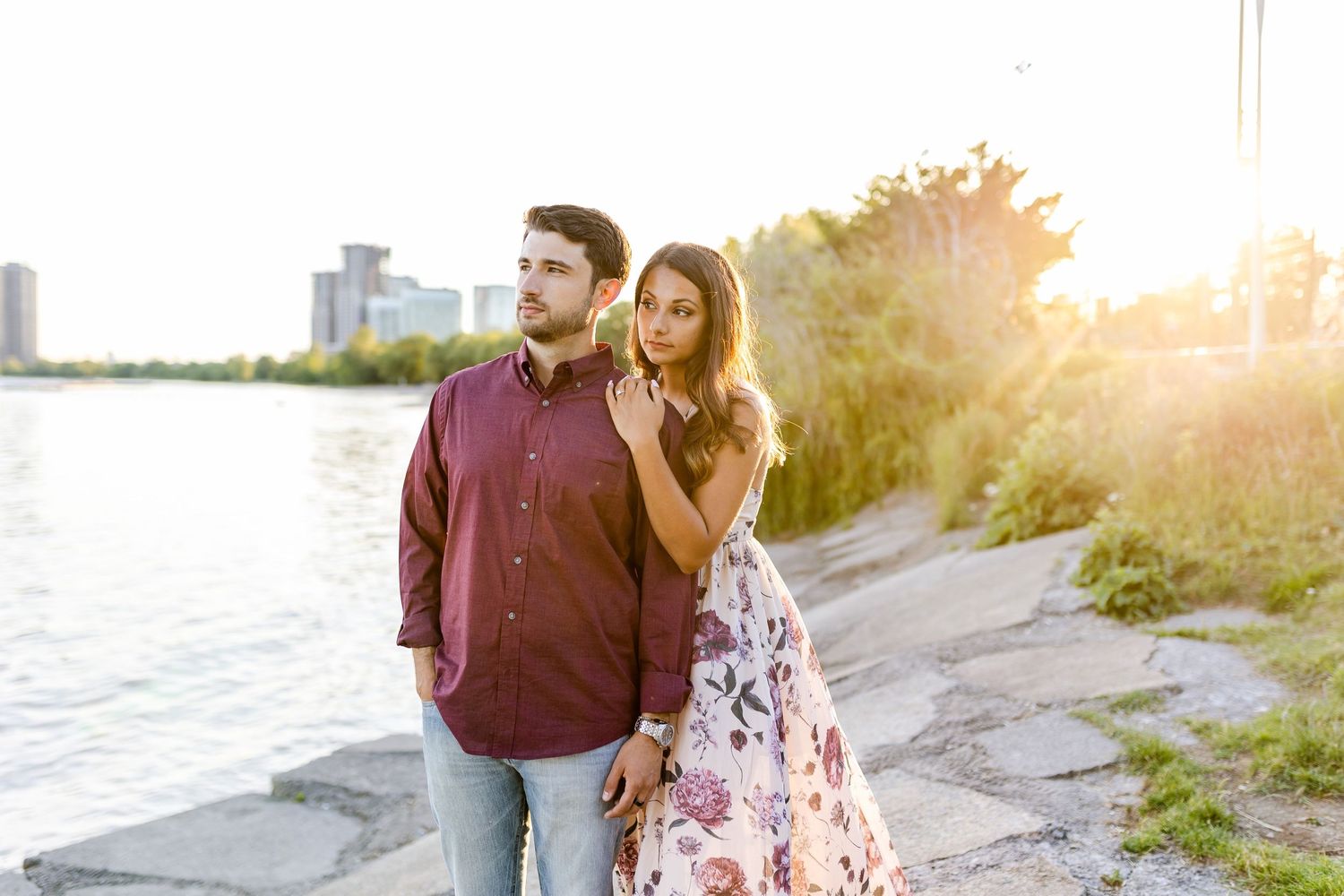 Couple in burgundy shirt and floral dress pose against sunset waterfront with city skyline.