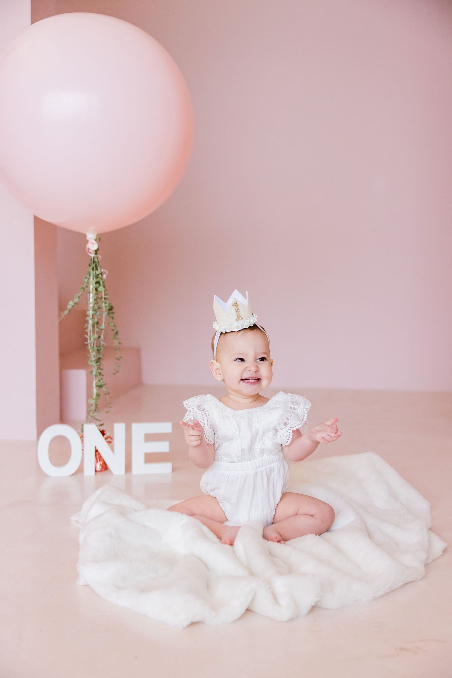 First birthday celebration setup with white dress, crown, and pink balloon on soft pink background.