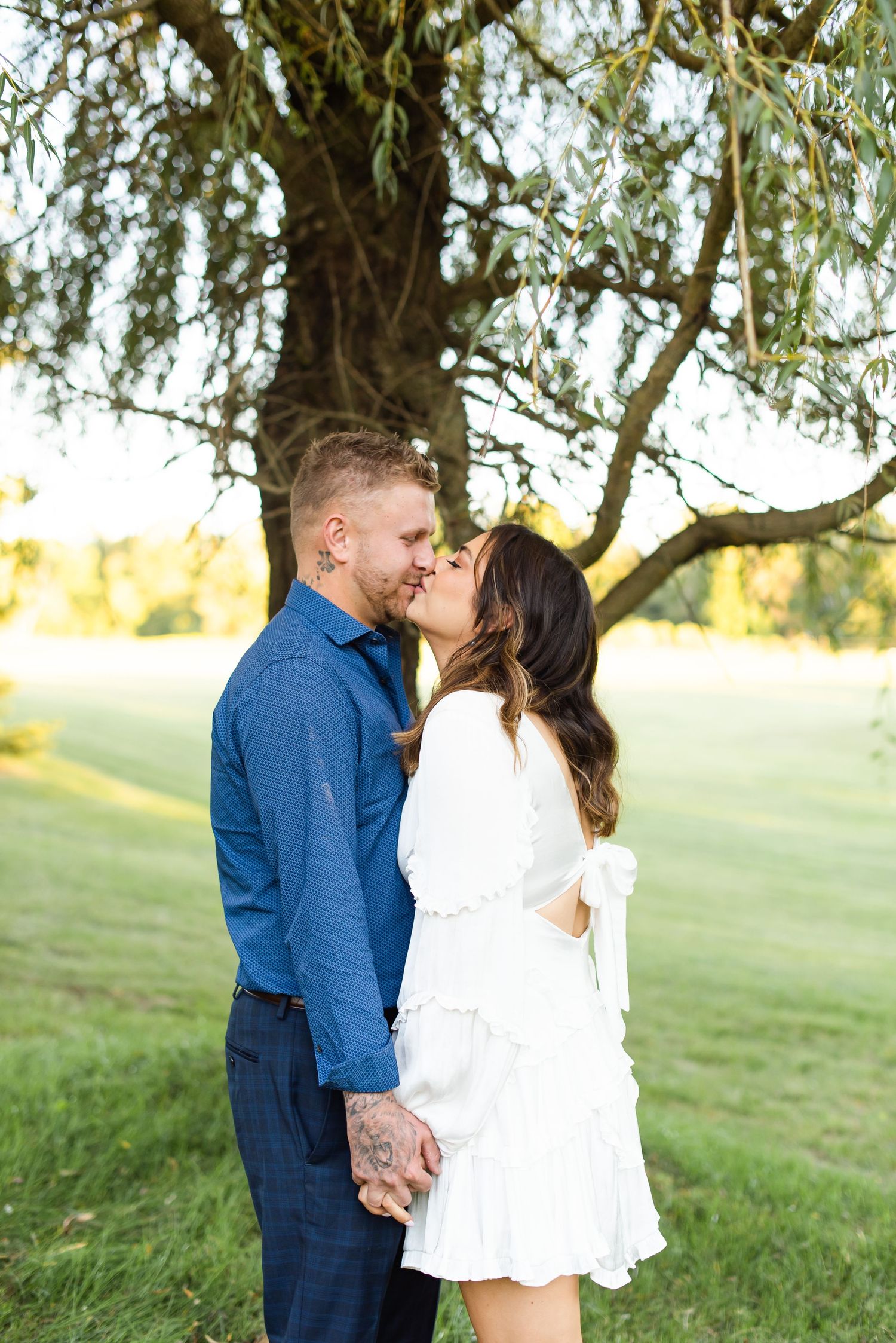 A romantic couple shares a tender moment under a weeping willow tree in golden sunlight.