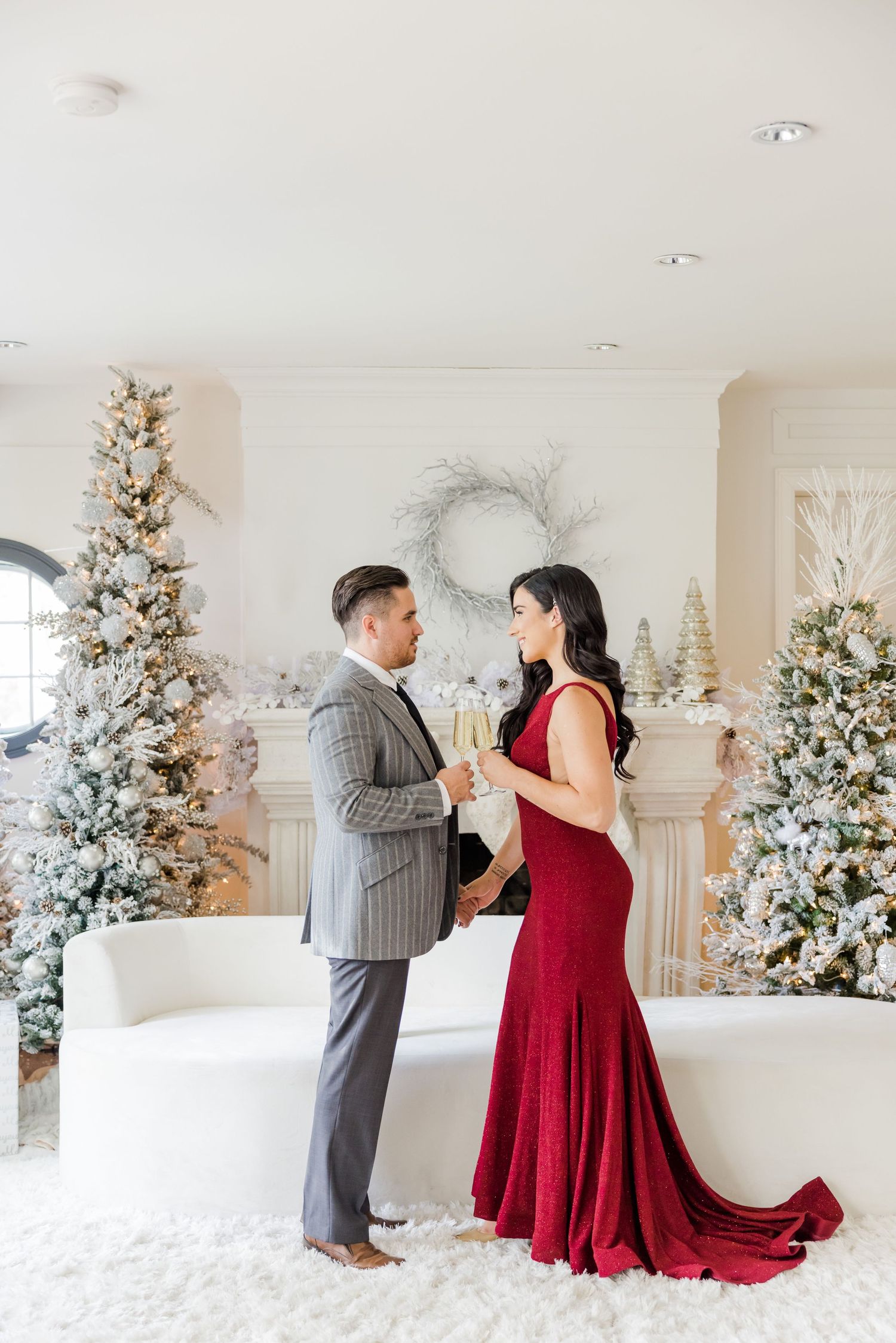 Couple in formal attire posing in front of decorated Christmas trees in an elegant white room.