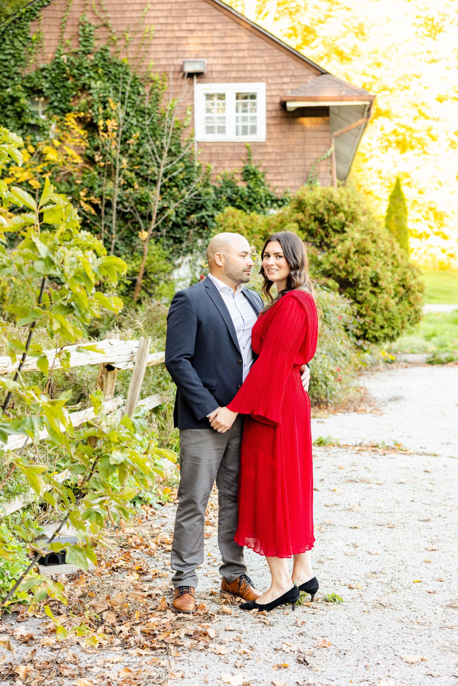 Elegant couple poses on garden path with brick house and autumn foliage in background.