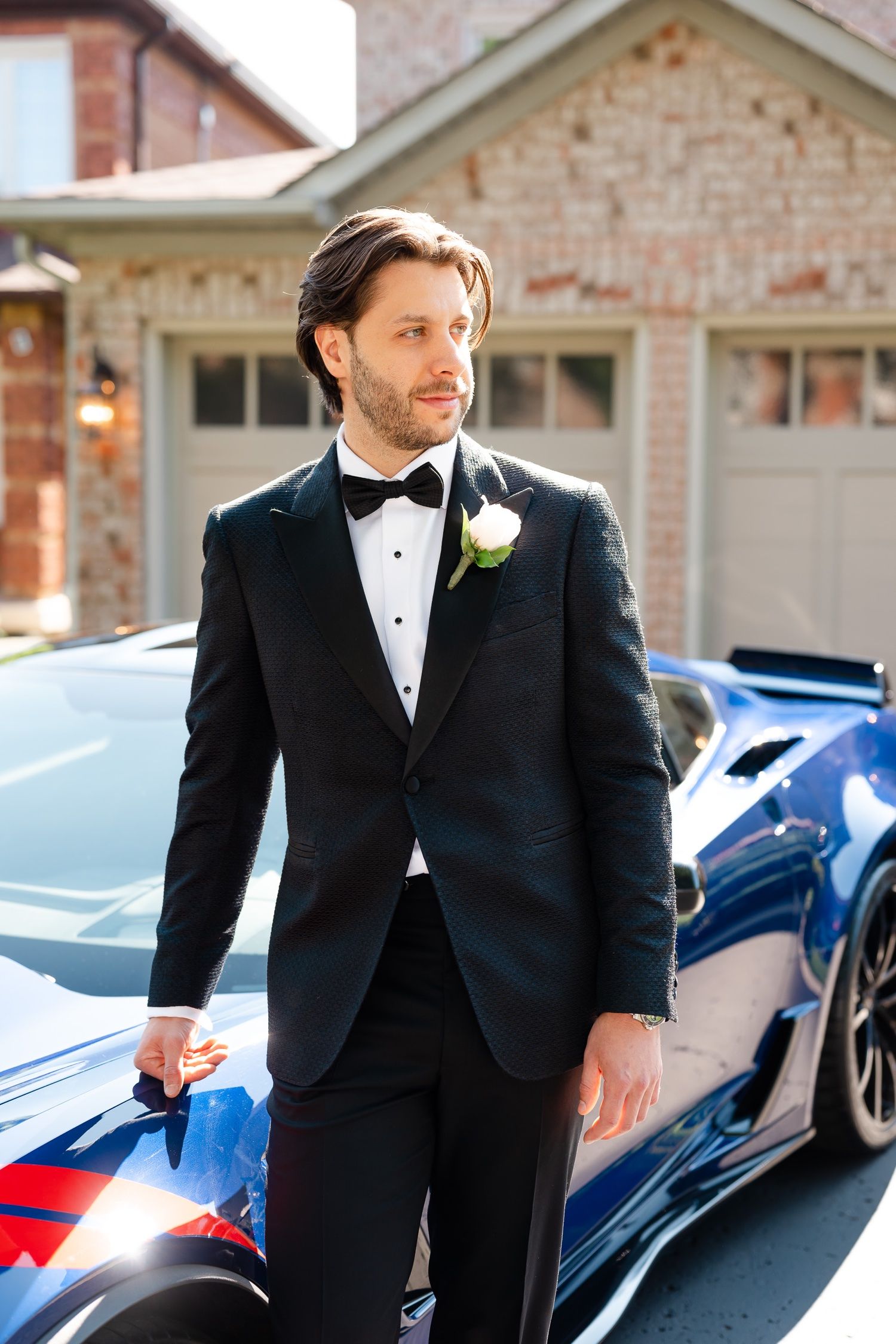 Formal portrait against blue sports car with boutonniere on black tuxedo.