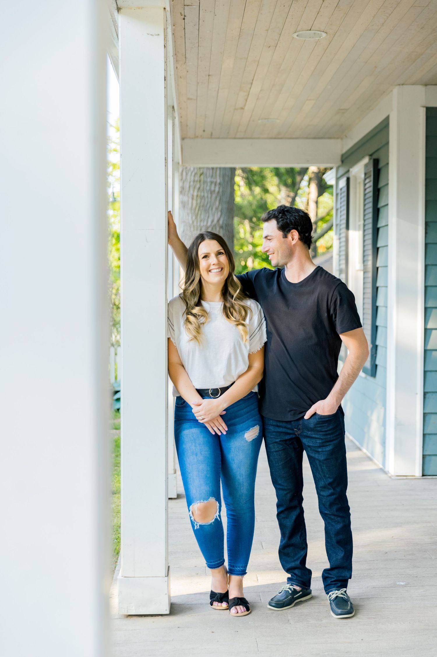 A couple poses casually on a covered porch with white columns and wooden ceiling.