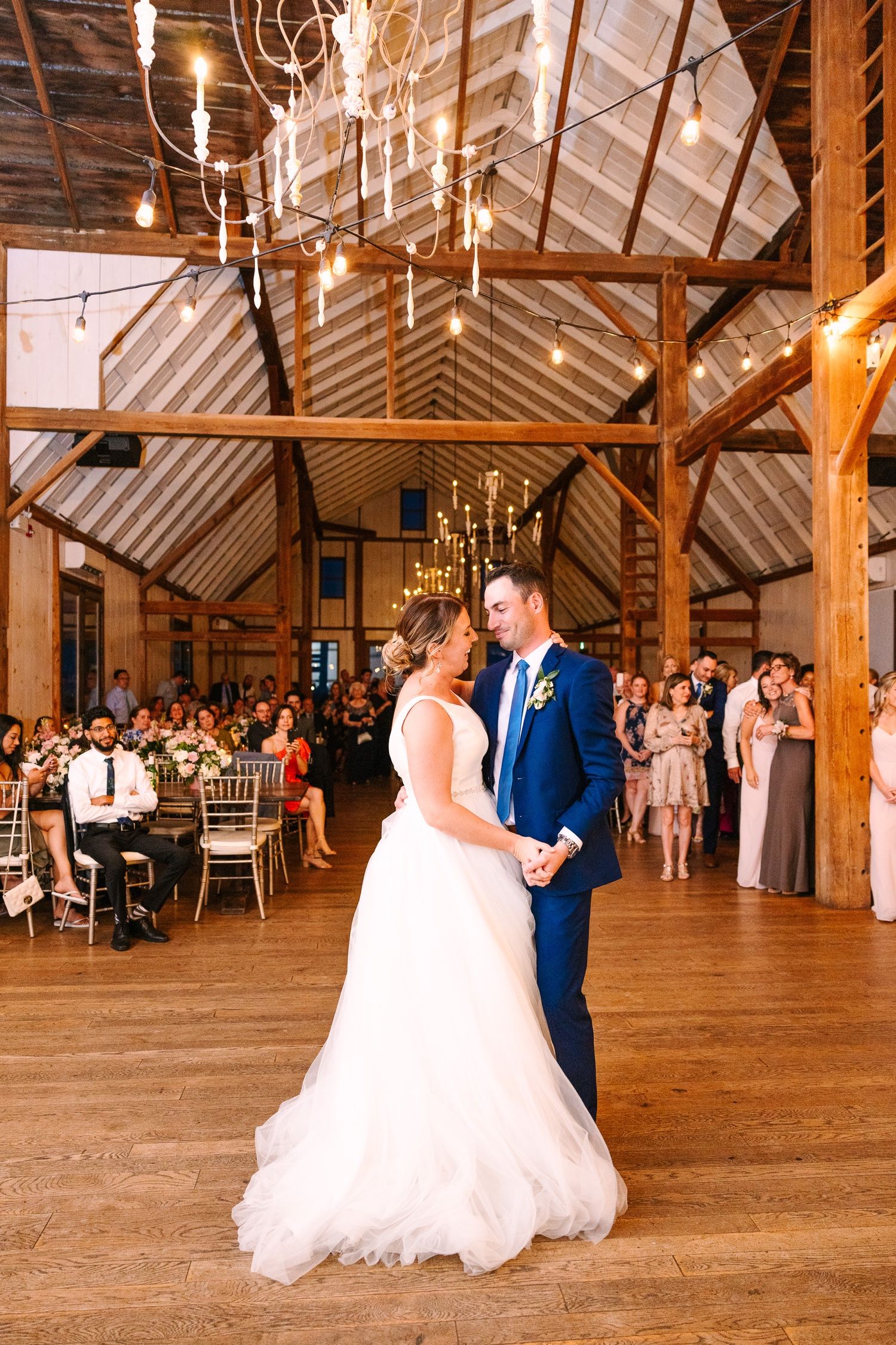 Romantic first dance under wooden beams and string lights at barn wedding reception.