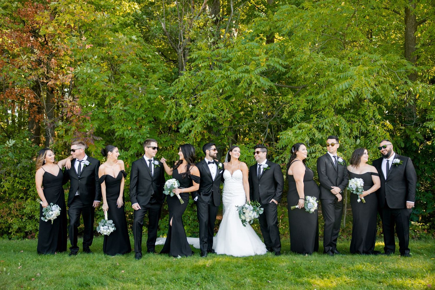 Wedding party in classic black attire poses casually on a lawn with fall foliage behind them.