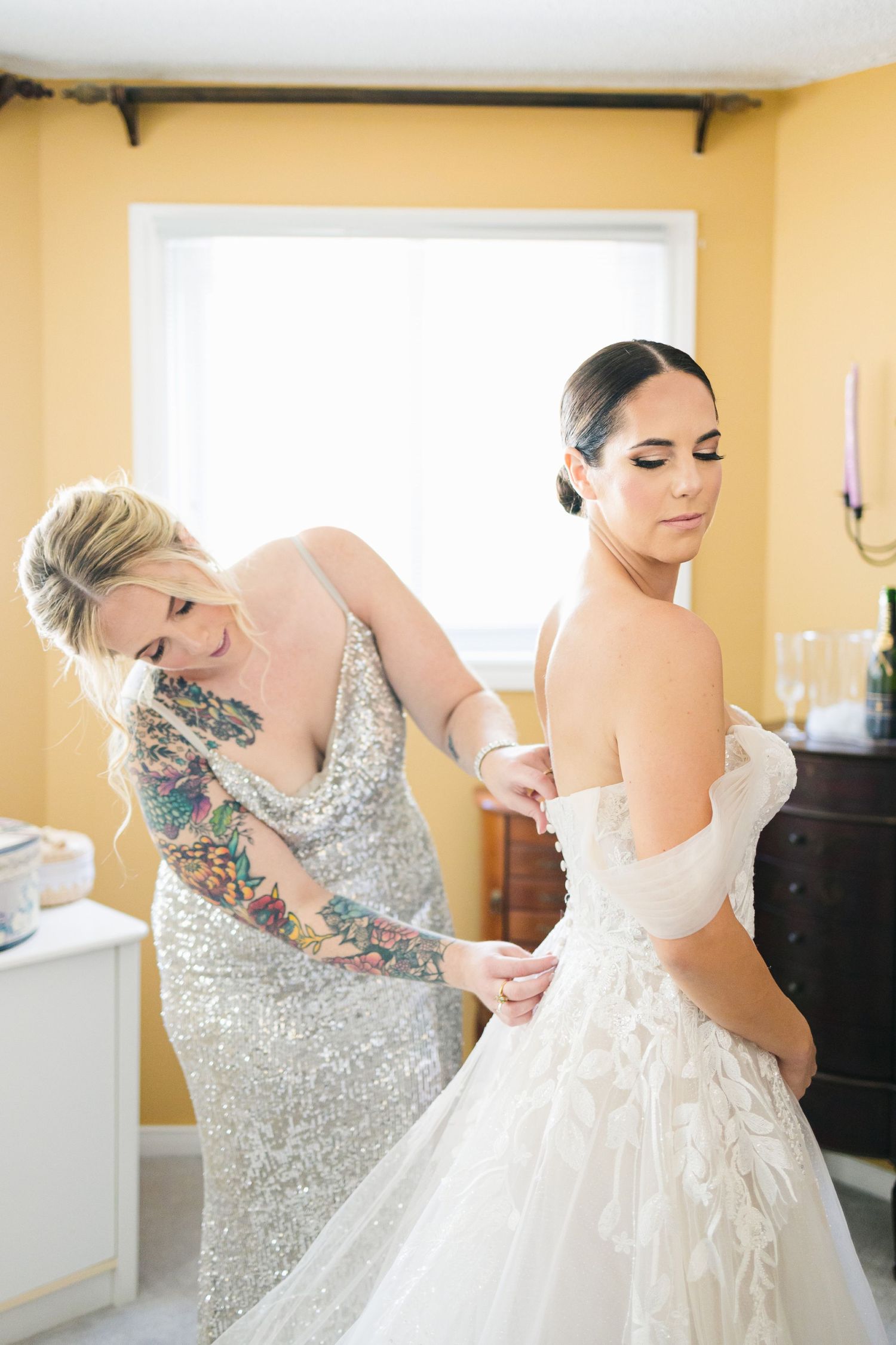 Behind the scenes wedding preparation moment in yellow room with elegant gowns.