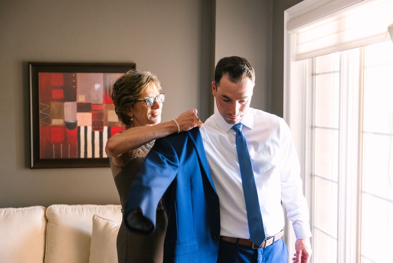 Wedding preparation scene with blue suit jacket being helped on in sunlit room.
