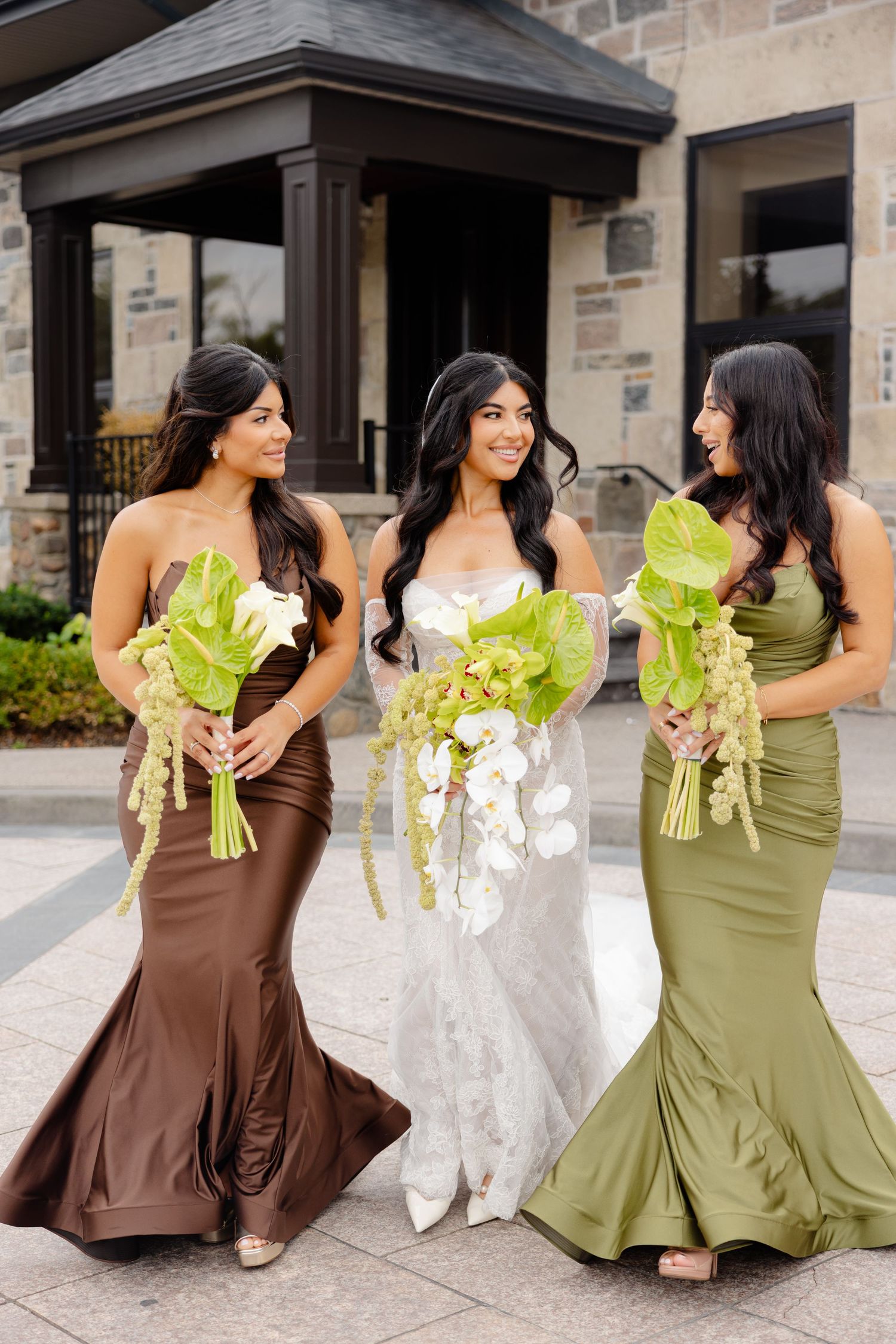 Wedding party in brown and olive green gowns holding fresh lettuce bouquets share candid laughs at venue entrance.