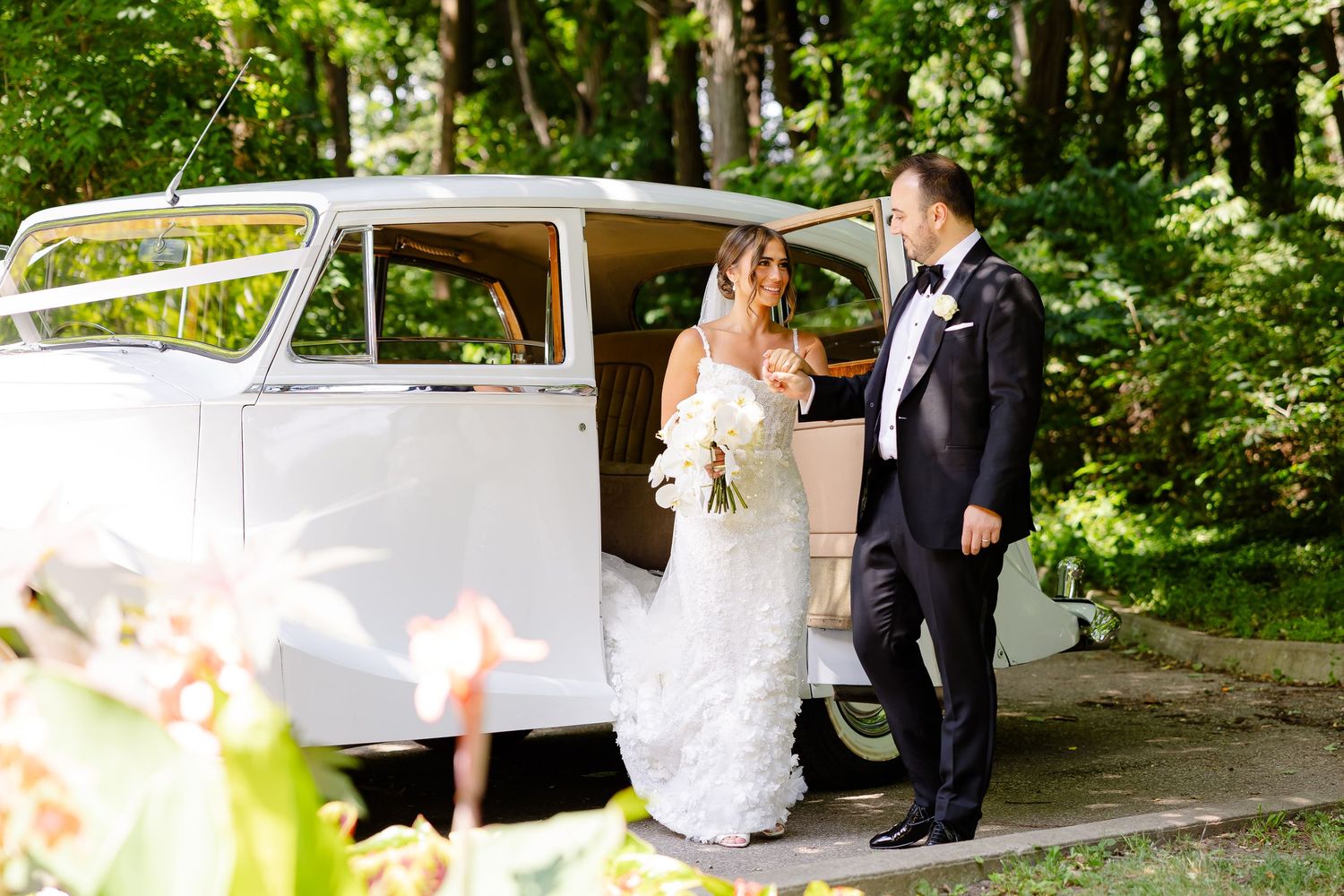 A couple in formal wedding attire stands beside a classic white vintage car in a lush green forest setting.