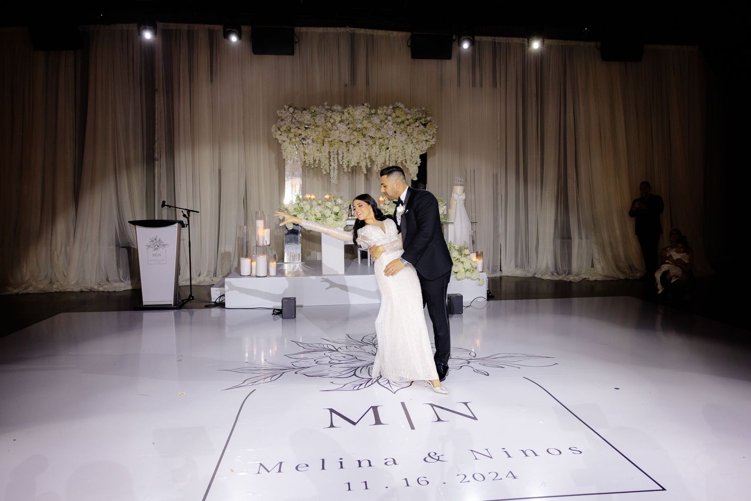 Celebratory dance moves on monogrammed floor with white floral arch and dramatic lighting.