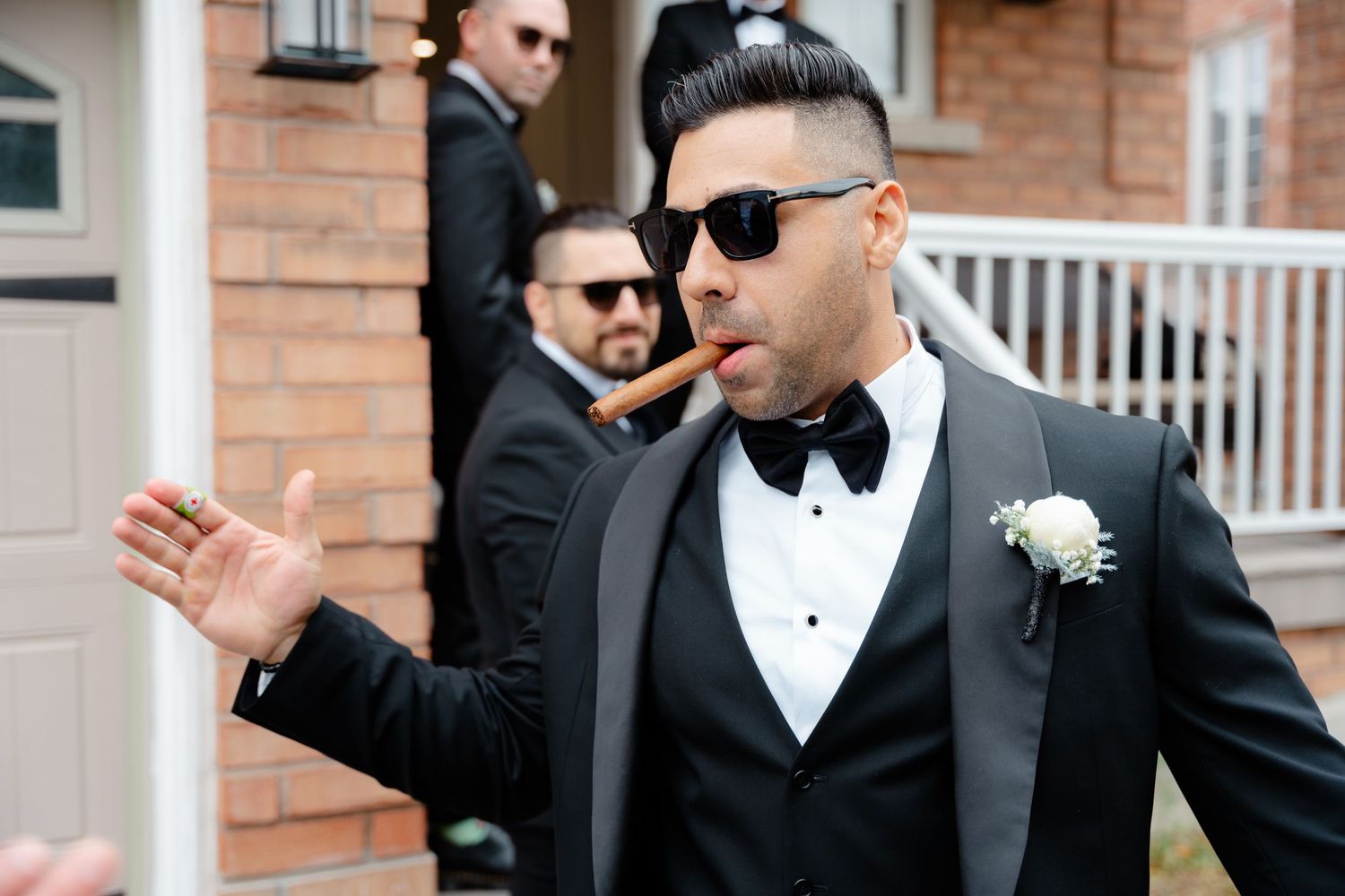 Groomsman in black tuxedo and sunglasses gesturing expressively outside brick building.